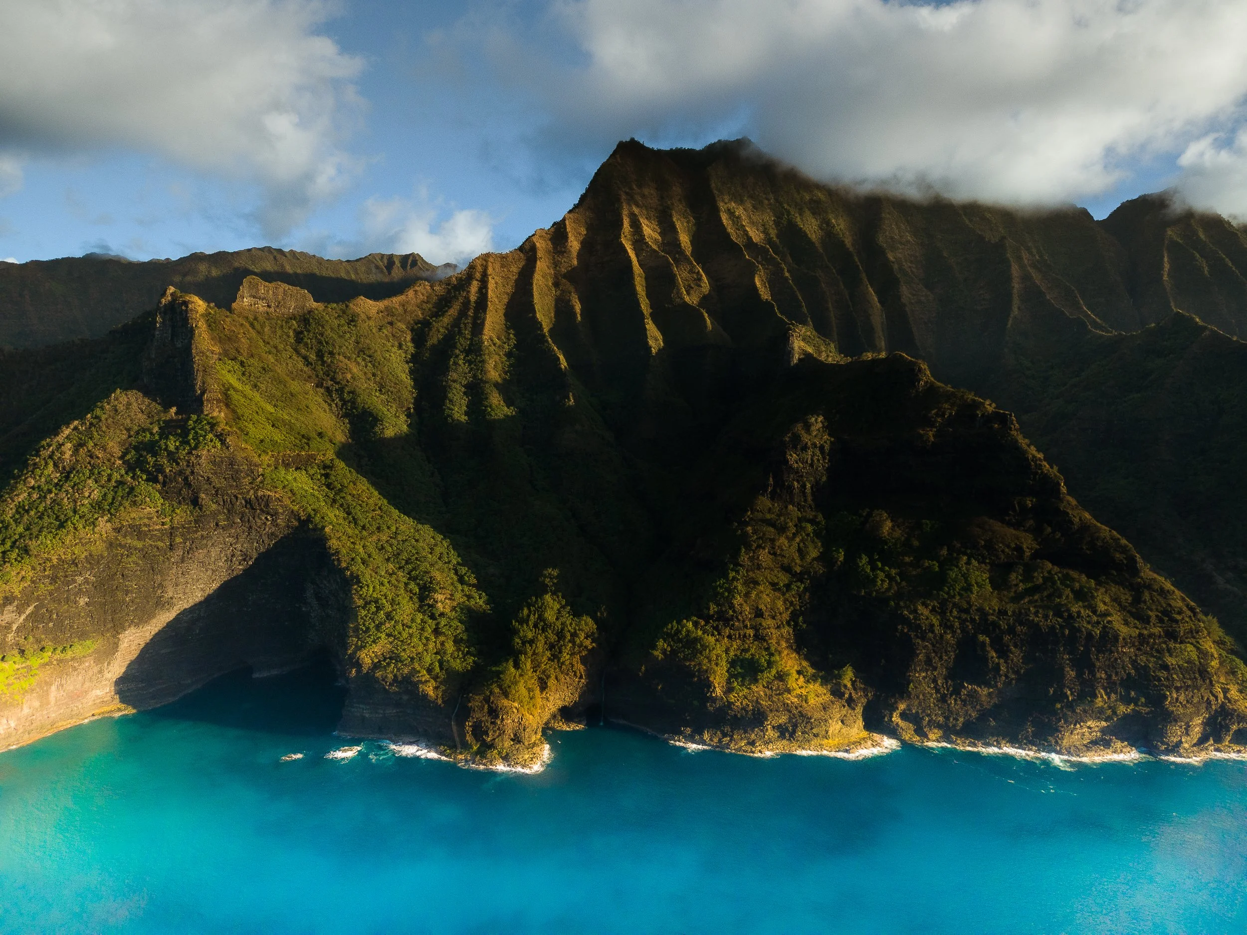 Aerial view of lush green mountains with steep cliffs meeting the blue ocean, partly covered by clouds.
