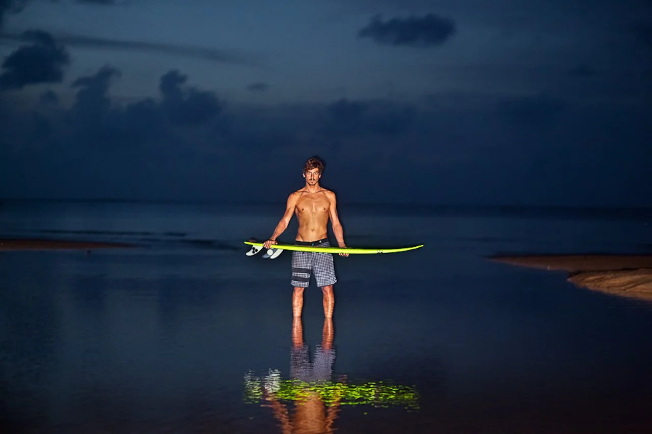A shirtless man holding a yellow surfboard stands in shallow water at night, with a cloudy sky and dark ocean in the background.