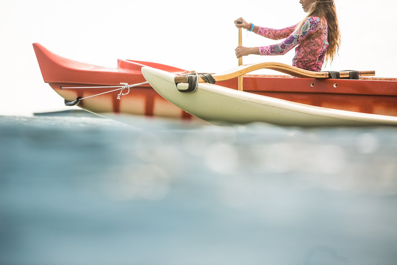 A person with long hair in a colorful long-sleeve shirt paddling a kayak on the water, with a second kayak partially visible next to them.