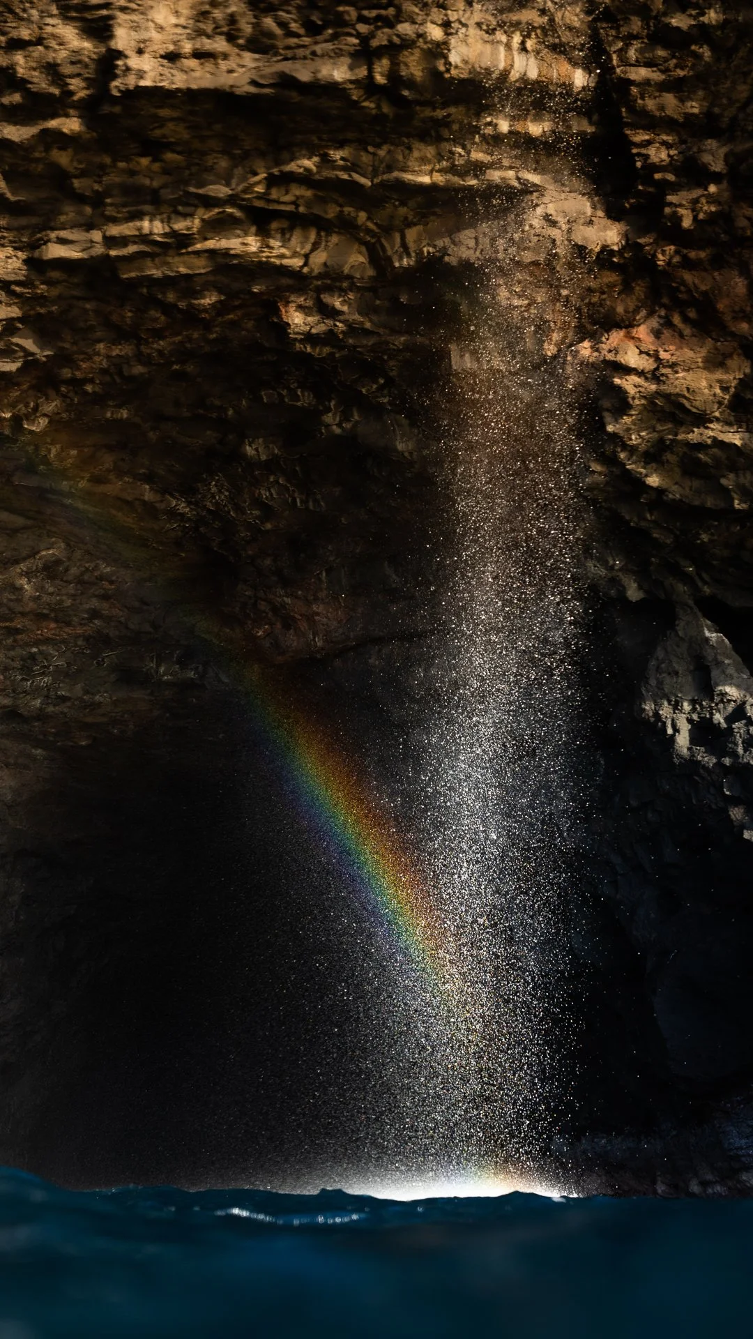 Waterfall inside a dark cave with water spray creating a rainbow effect.