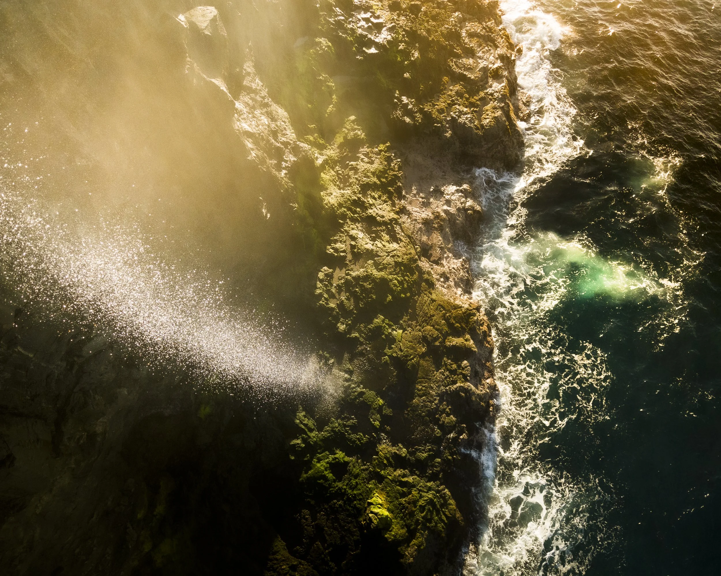 An aerial view of ocean waves crashing against rocks on a coastline with sunlight shining over the water.