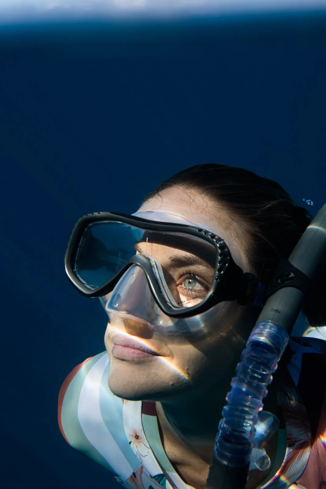 A woman with green eyes submerged underwater, wearing a diving mask and snorkel, looking upwards with sunlight reflecting on her face.