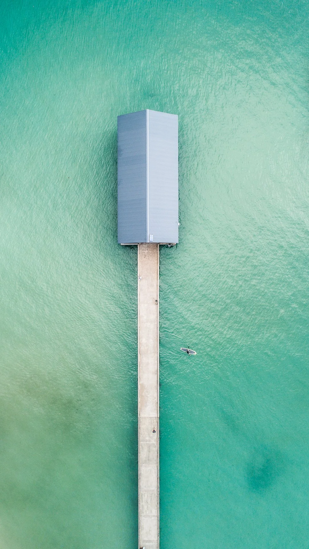 An aerial view of a long pier extending into turquoise water, with a rectangular building at the end of the pier and a person paddleboarding nearby.