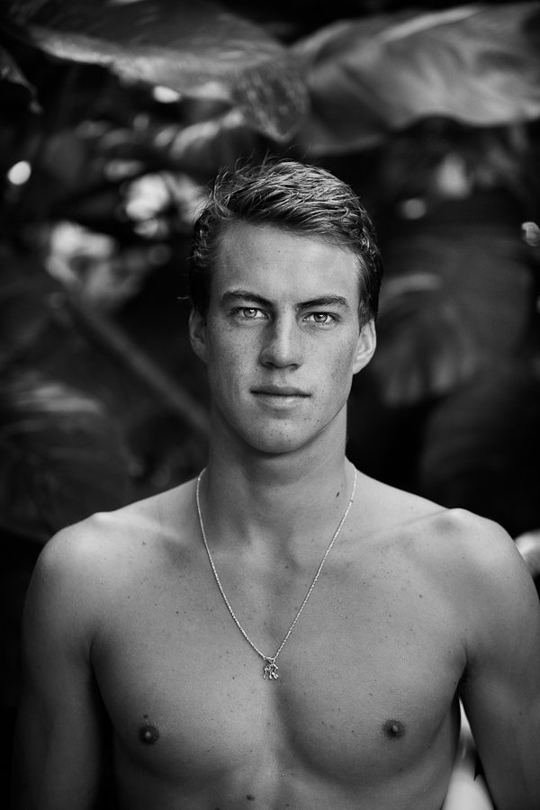 Black and white portrait of a young shirtless man with short hair, wearing a necklace, standing in front of large leaves.