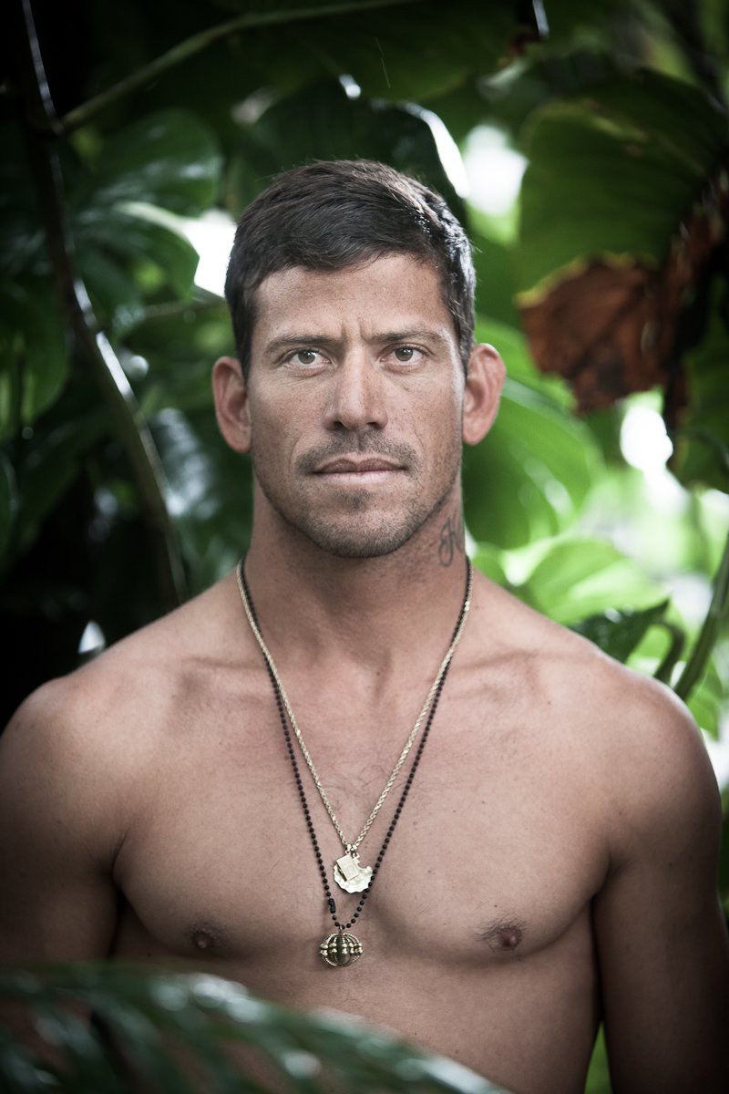 A shirtless man with short dark hair and light skin standing among large green leaves, looking directly at the camera with a serious expression.