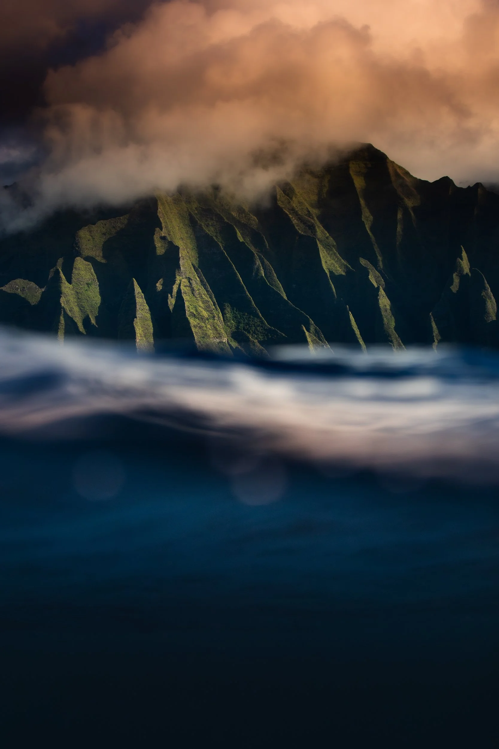 Mountain cliffs rising from the ocean with clouds above, illuminated by sunset light.