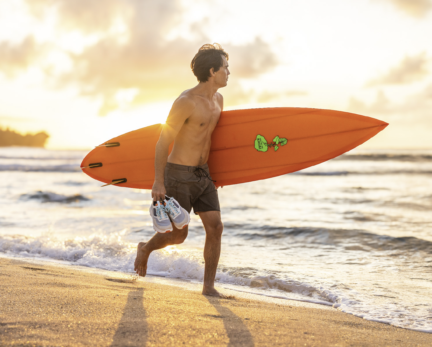 A man with dark hair carrying a surfboard and shoes on a beach at sunset.