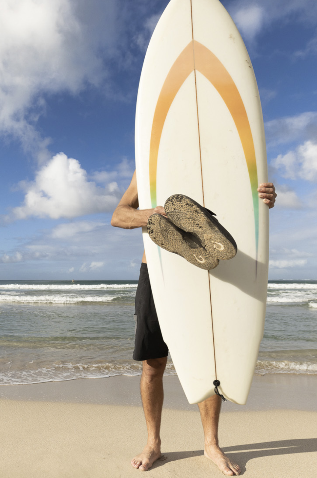 a person standing on the beach, holding a surfboard with flip-flops hanging on the front, facing the ocean.