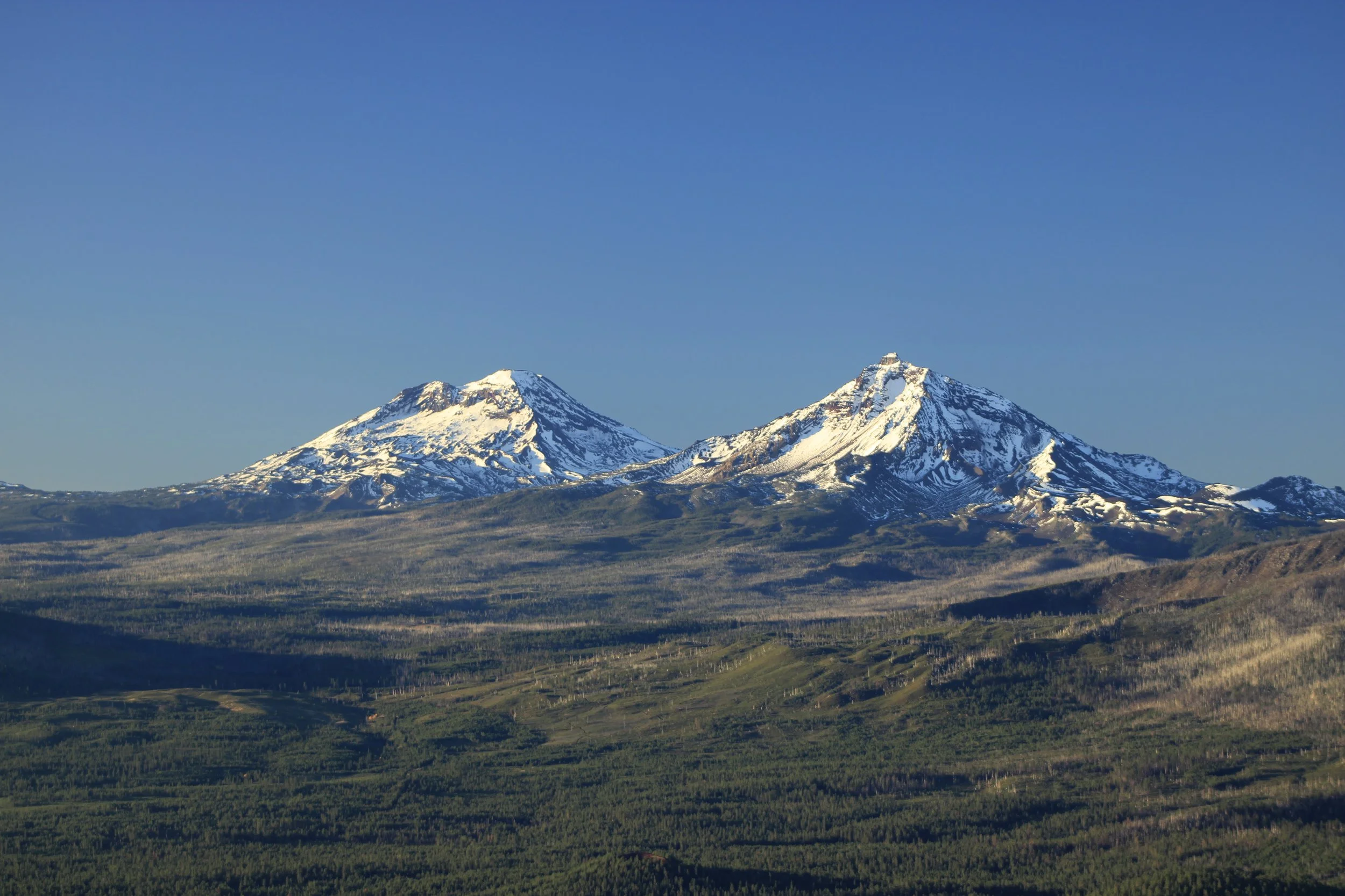 Two snow-capped volcanic mountains above a green landscape under a clear blue sky.