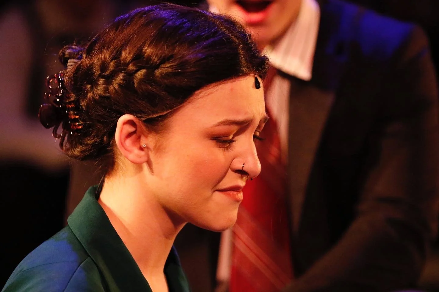 A young woman with dark red hair styled in a braided updo, closed eyes, and a gentle smile, wearing a dark green jacket, with a nose piercing and small earrings, in a dimly lit indoor setting.