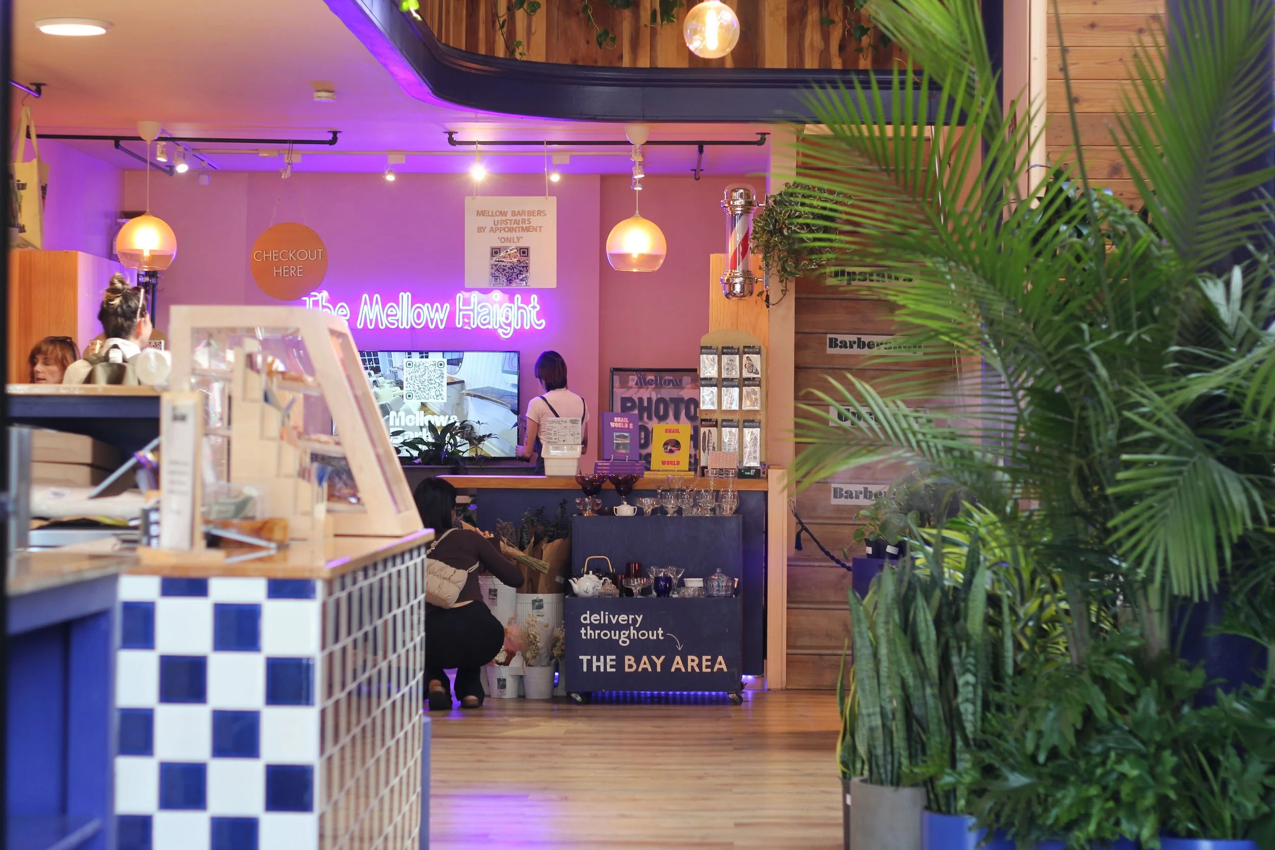 Interior of a cafe or restaurant with a neon sign that reads 'The Mellow Height,' a counter with a staff member, and lush green plants in the foreground.