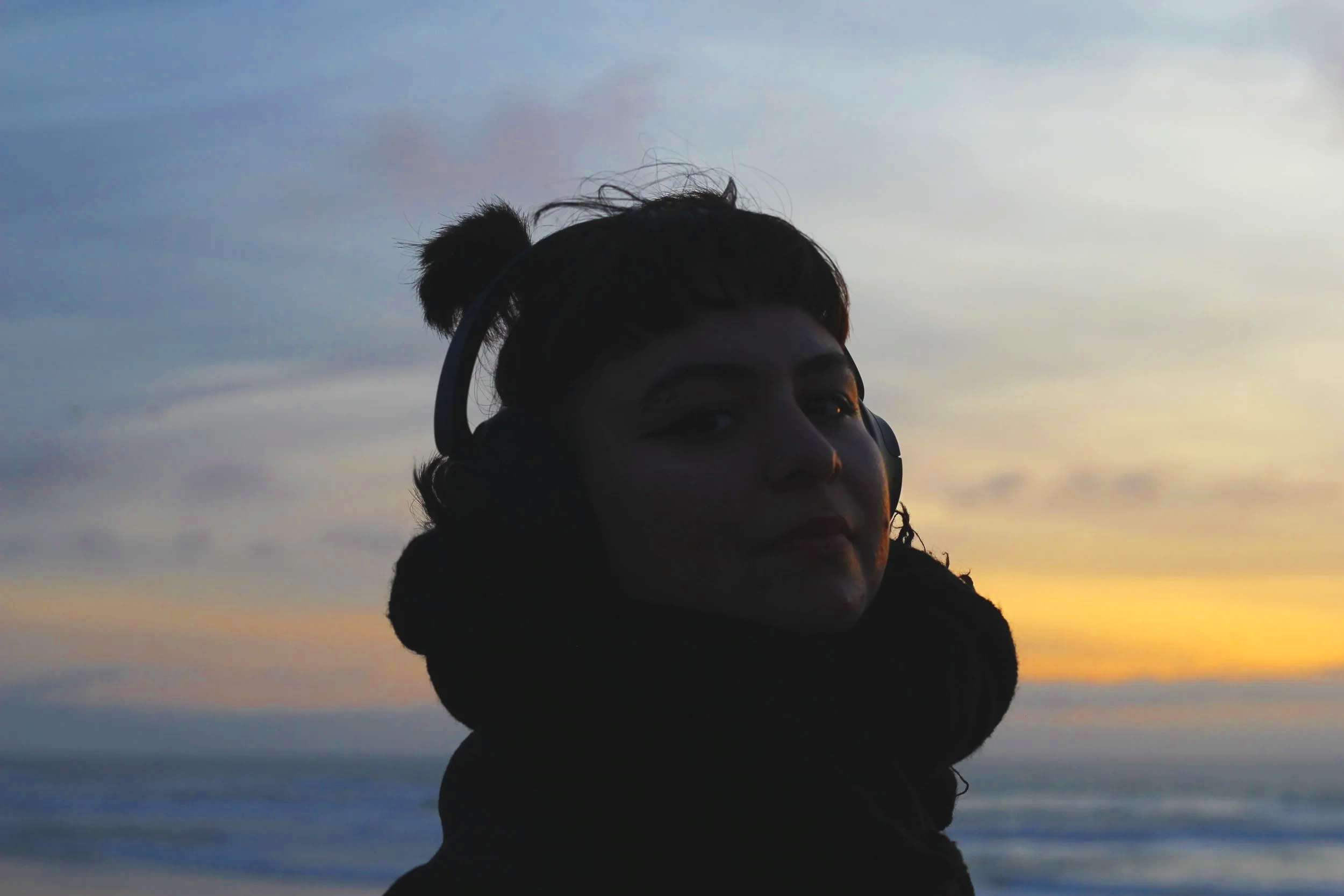 A woman with dark hair styled in a bun, wearing headphones and a black scarf, is outdoors during sunset with the sky and ocean in the background.