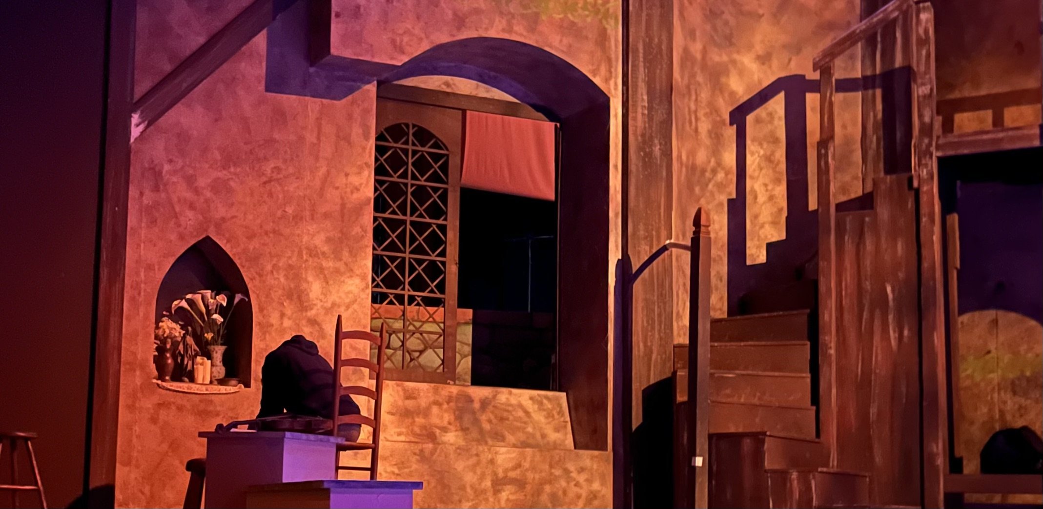 Stage set designed to resemble a rustic house or castle interior with wooden stairs, a chair, a small table with flowers, and walls with a textured warm-colored finish.