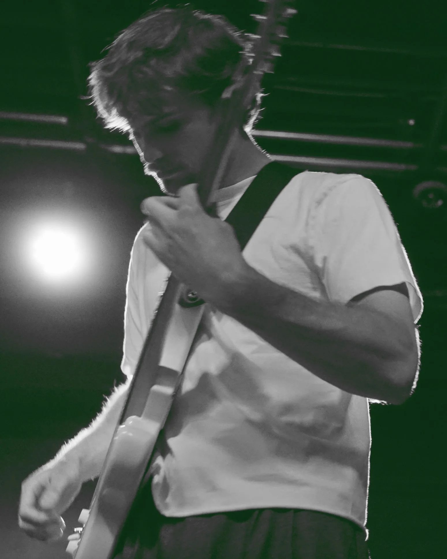 A young man playing an electric guitar on stage, wearing a white t-shirt and a guitar strap, with stage lights in the background.