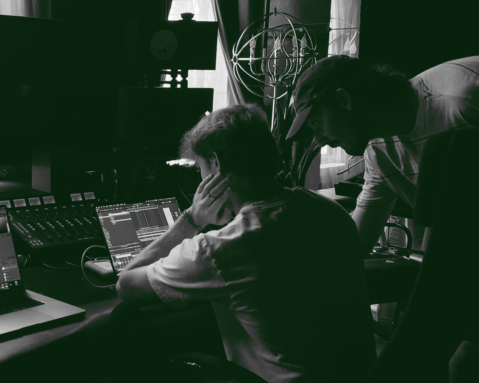 Two individuals working together in a music studio, one seated at a laptop and mixing console, the other standing and providing assistance, with musical equipment and a chandelier in the background.