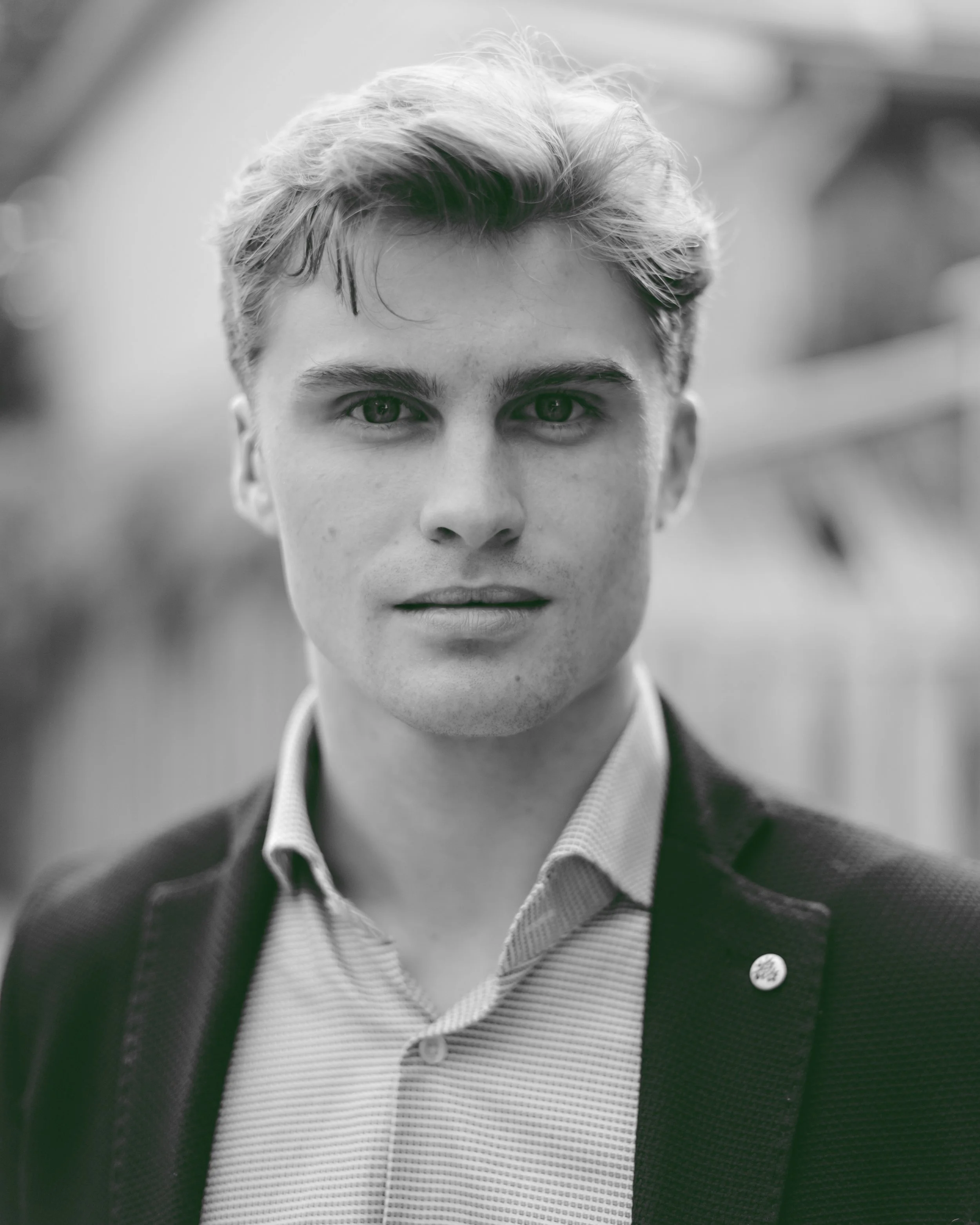 Black and white close-up portrait of a young man with light hair, wearing a collared shirt and jacket, looking directly at the camera with a neutral expression.