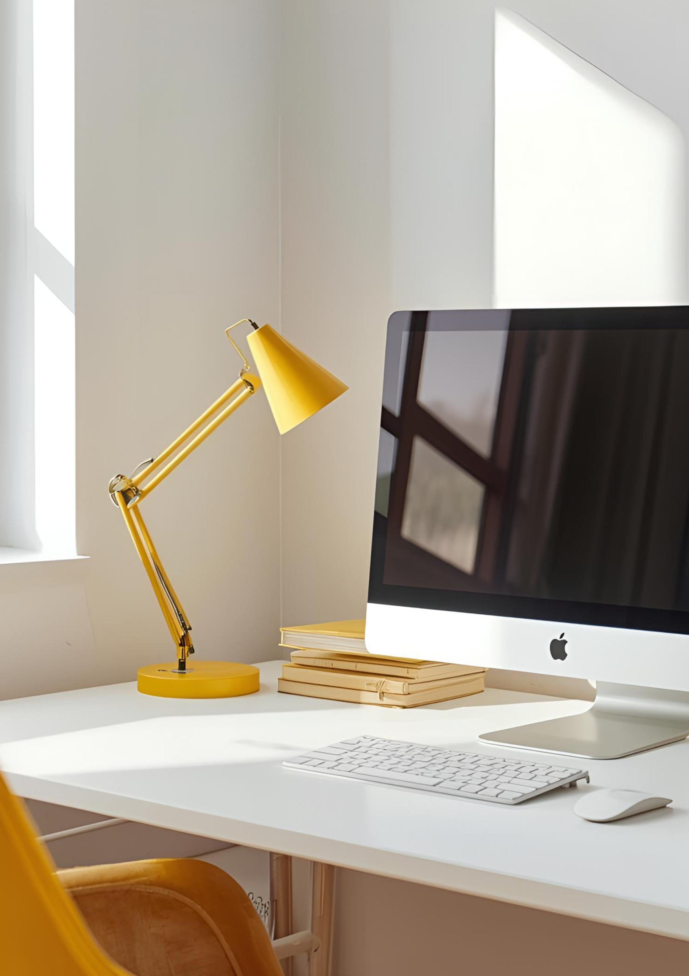 Bureau blanc avec un ordinateur iMac, un clavier et une souris, une lampe de bureau jaune et une pile de livres ou Carnets jaunes près d'une fenêtre.
