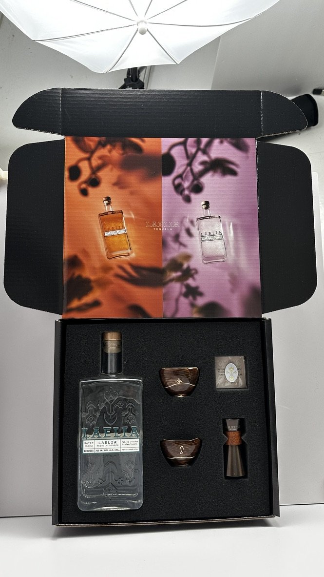 Open black box containing a bottle of Tequila Azul, two small bowls, a cork, and a small metal tool, with a colorful card showing two bottles, one amber and one clear, behind the box. There is a white umbrella overhead.