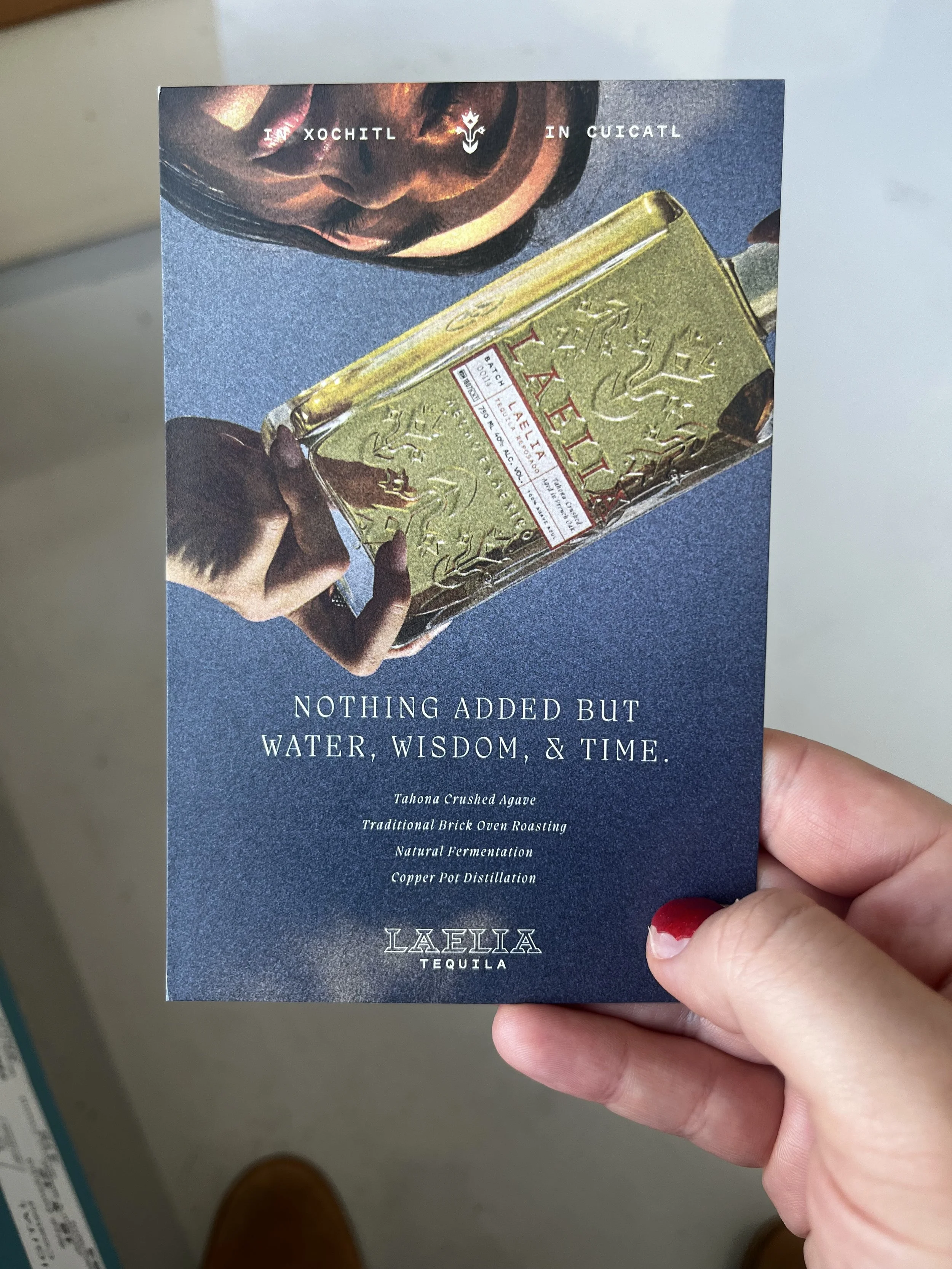 A person holding a dark blue card with an image of a hand holding a bottle of Laelia tequila. The text on the card reads, 'Nothing added but water, wisdom, & time. Tahona crushed agave, traditional brick oven roasting, natural fermentation, copper po
