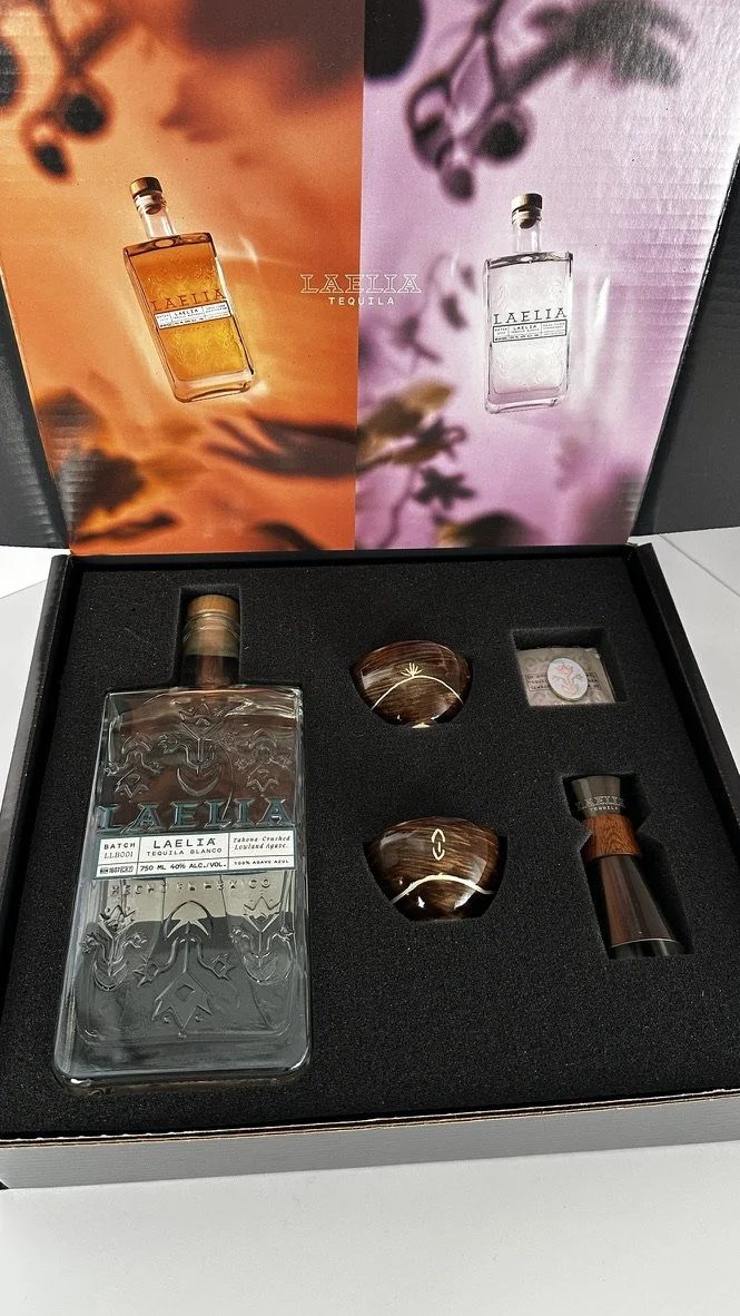A tequila gift set with a bottle, two cups, a small container, and a wooden-handled tool inside a foam insert, with colorful Laelia tequila branding in the background.