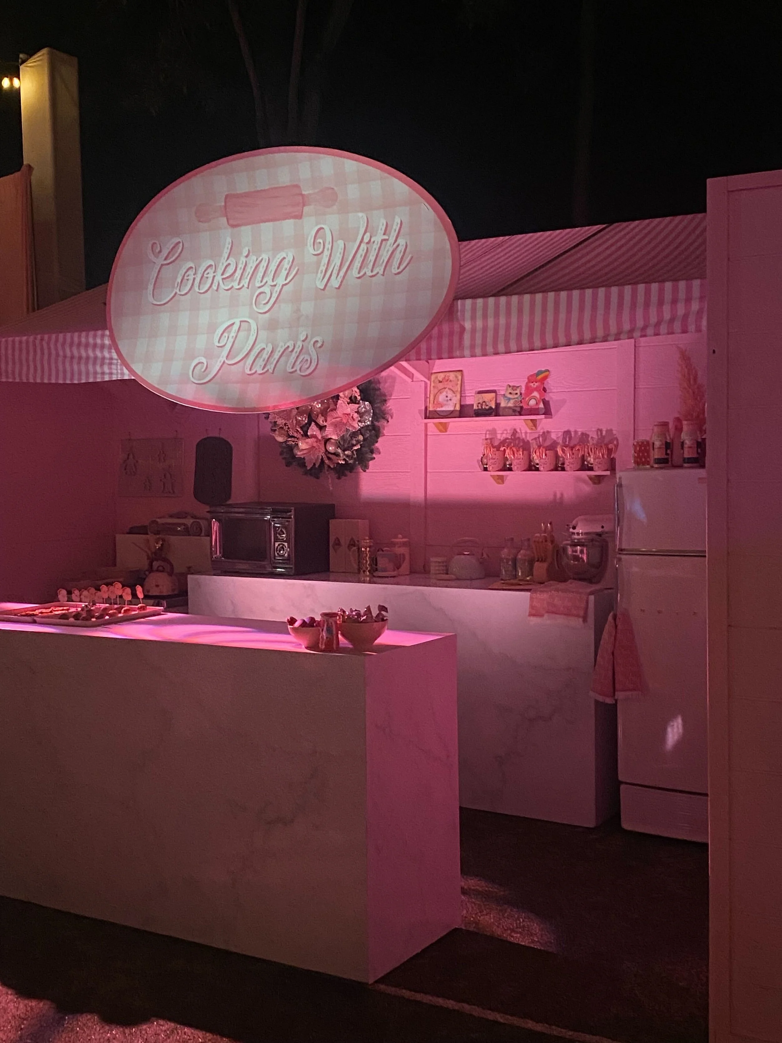 A pink-themed kitchen display at an event with a sign that reads 'Cooking With Paris', decorated with cupcakes, bowls, and kitchen appliances.