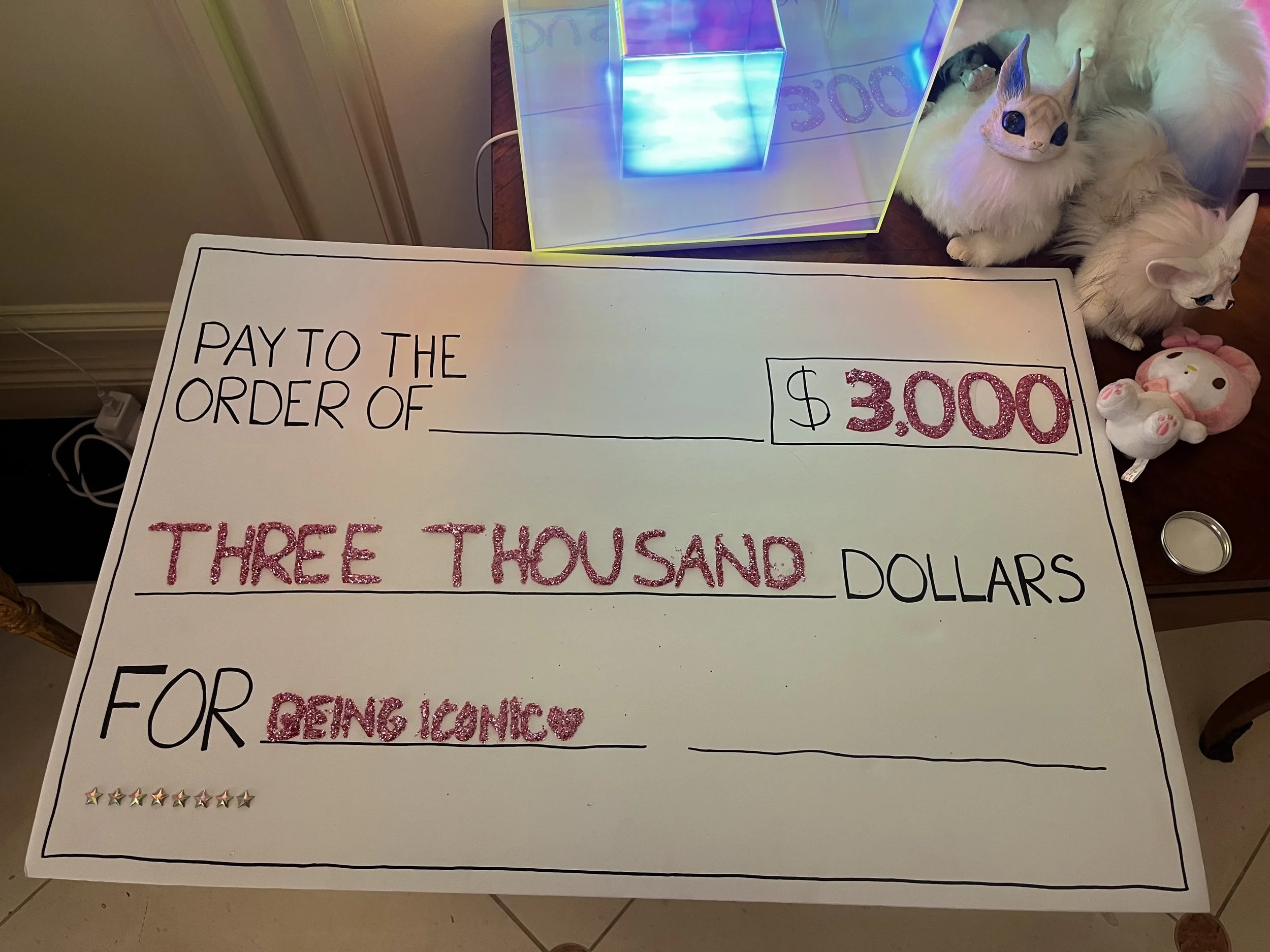 A large, jewelry-style check with black and pink glitter accents, reading: 'Pay to the order of ___ $3000 Three thousand dollars for being iconic.' The check is on a wooden table next to plush toys, including two white cats, a unicorn, and a pink Hel