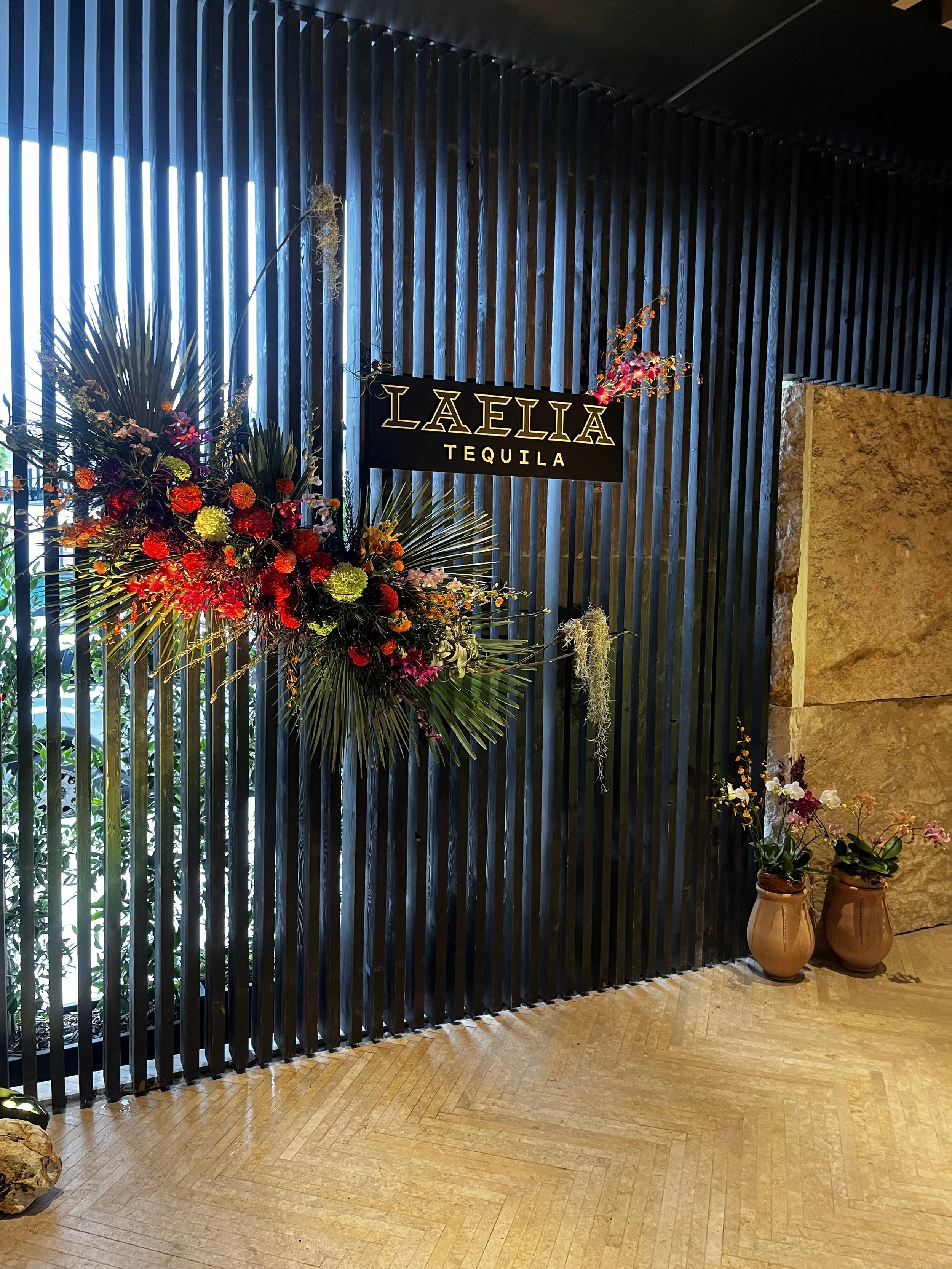 Decorative floral arrangement with red, pink, and yellow flowers on a black slatted wall, with a "Laelia Tequila" sign above it, and two potted orchids in clay pots on the floor.