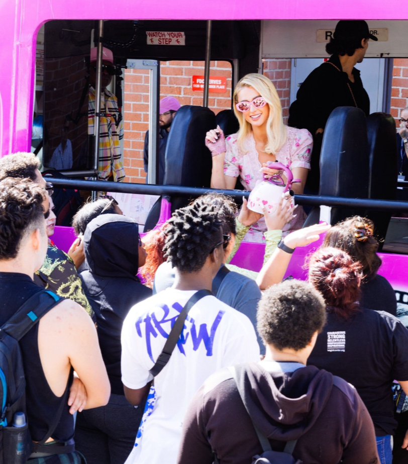 A woman with blonde hair, pink sunglasses, and a pink floral dress is waving and smiling from a pink bus window, greeting a crowd of people standing outside.