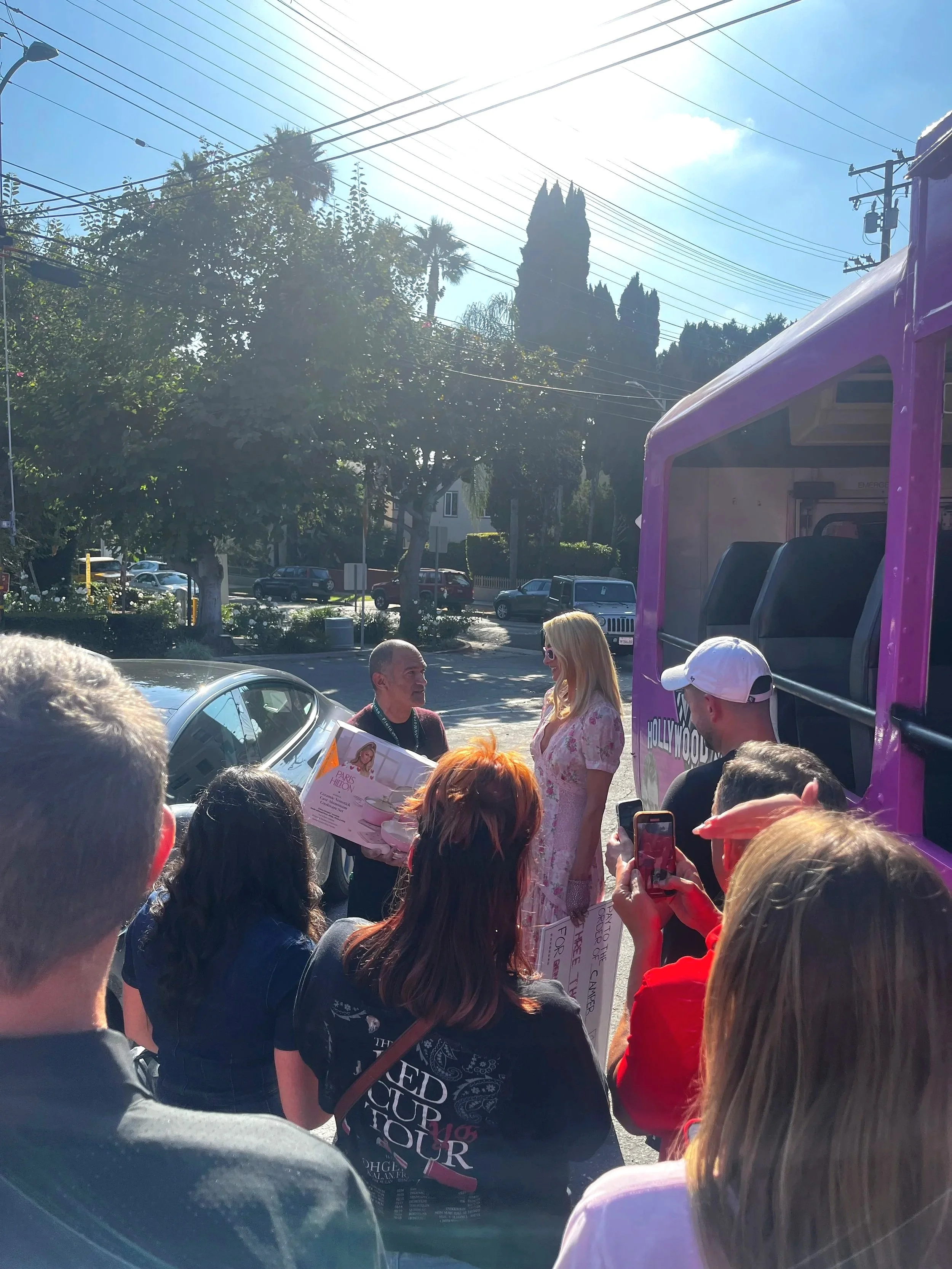A group of people gathered outdoors on a sunny day, with one man presenting a boxed item to a woman in a pink dress. The scene is near a purple vehicle, and there are trees, parked cars, and power lines in the background.