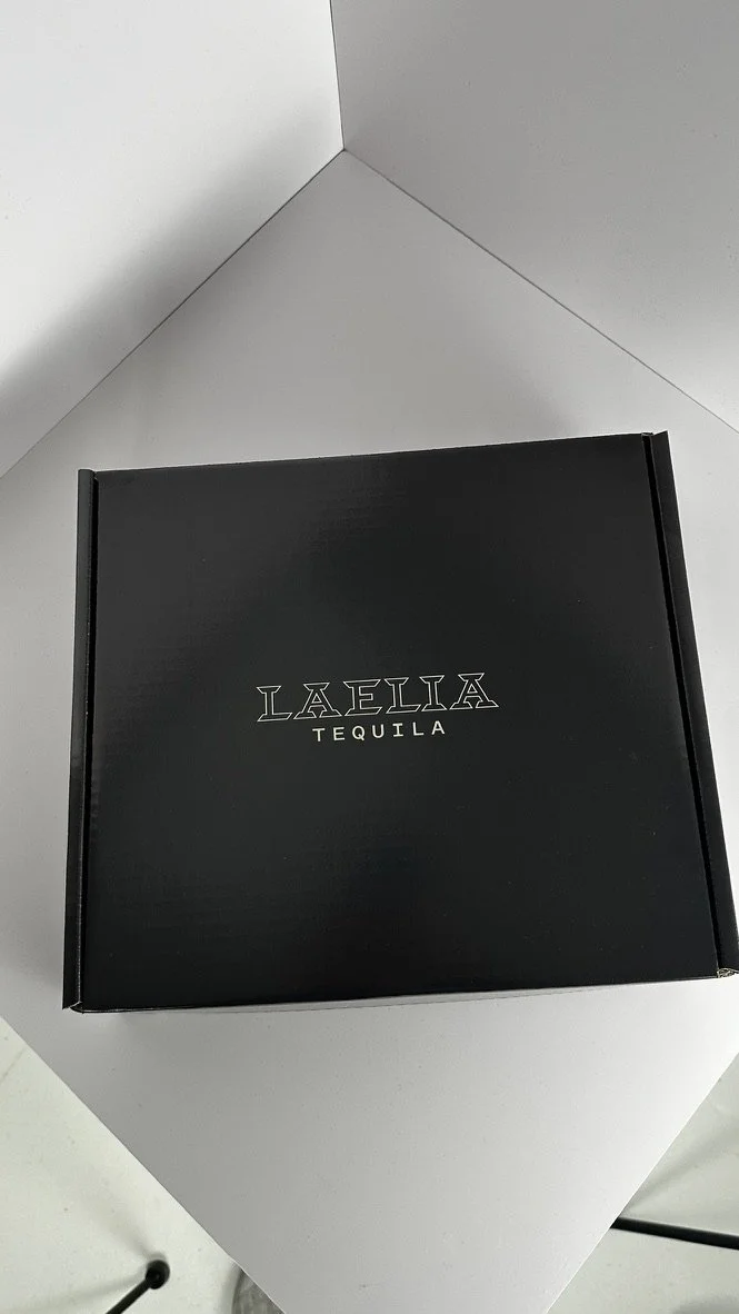 Black box with white text that reads 'LALIA TEQUILA' placed on a white surface against a white and gray corner wall.