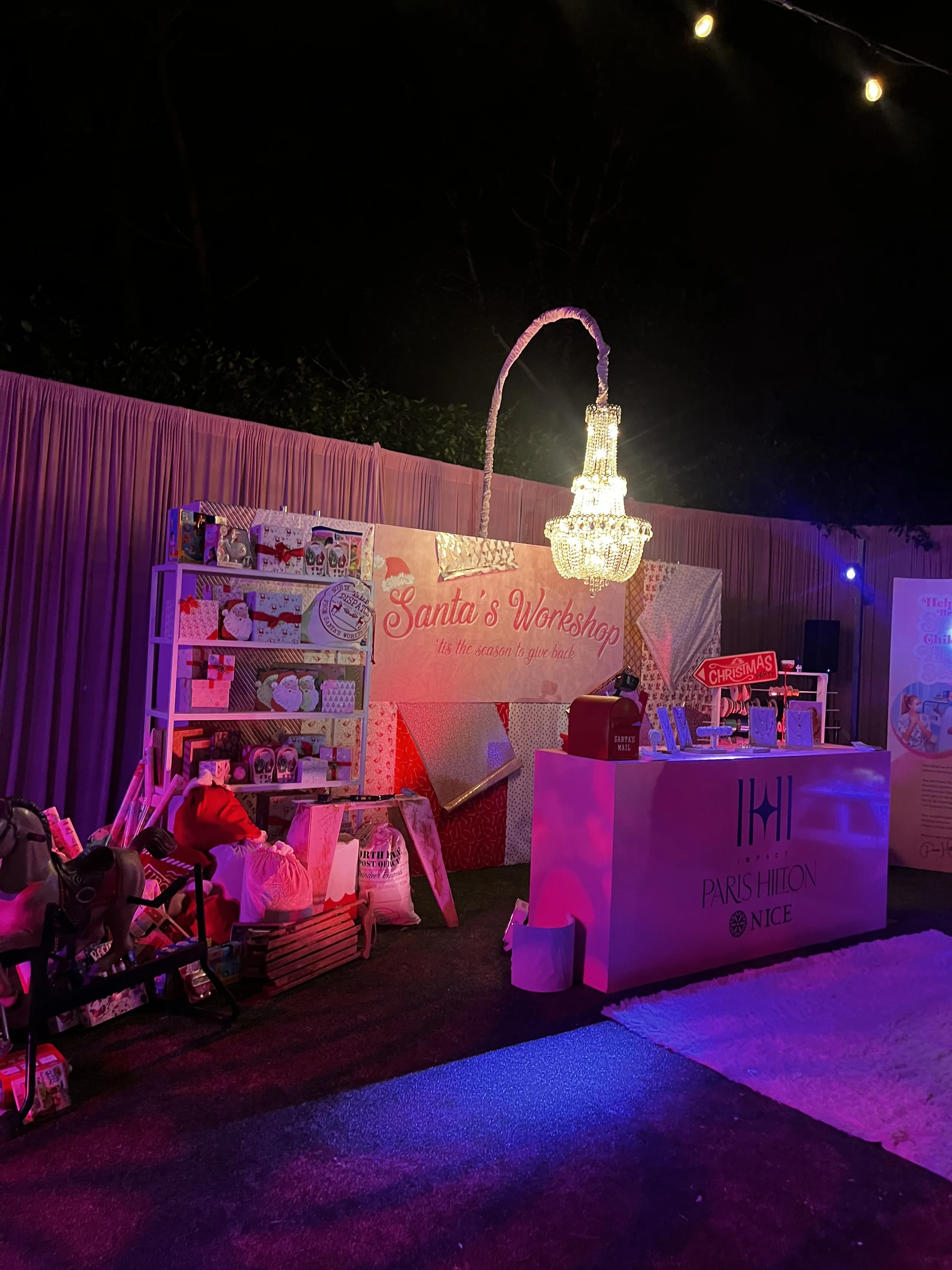 Decorative Christmas workshop setup with a backdrop reading 'Santa's Workshop'. Pink and red holiday-themed decorations, a chandelier, and a Paris Hilton-branded display counter with a 'Christmas' sign. Items like presents, a toy horse, and gift bags