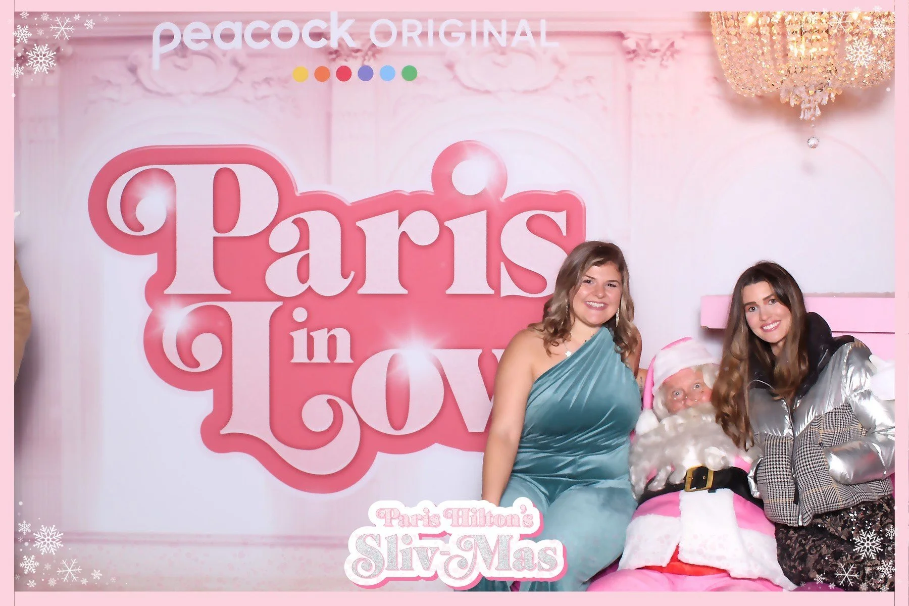 Two women posing with Santa Claus dressed in pink at a 'Paris in Love' themed event with pink and white decor, chandelier, and snowflake accents.