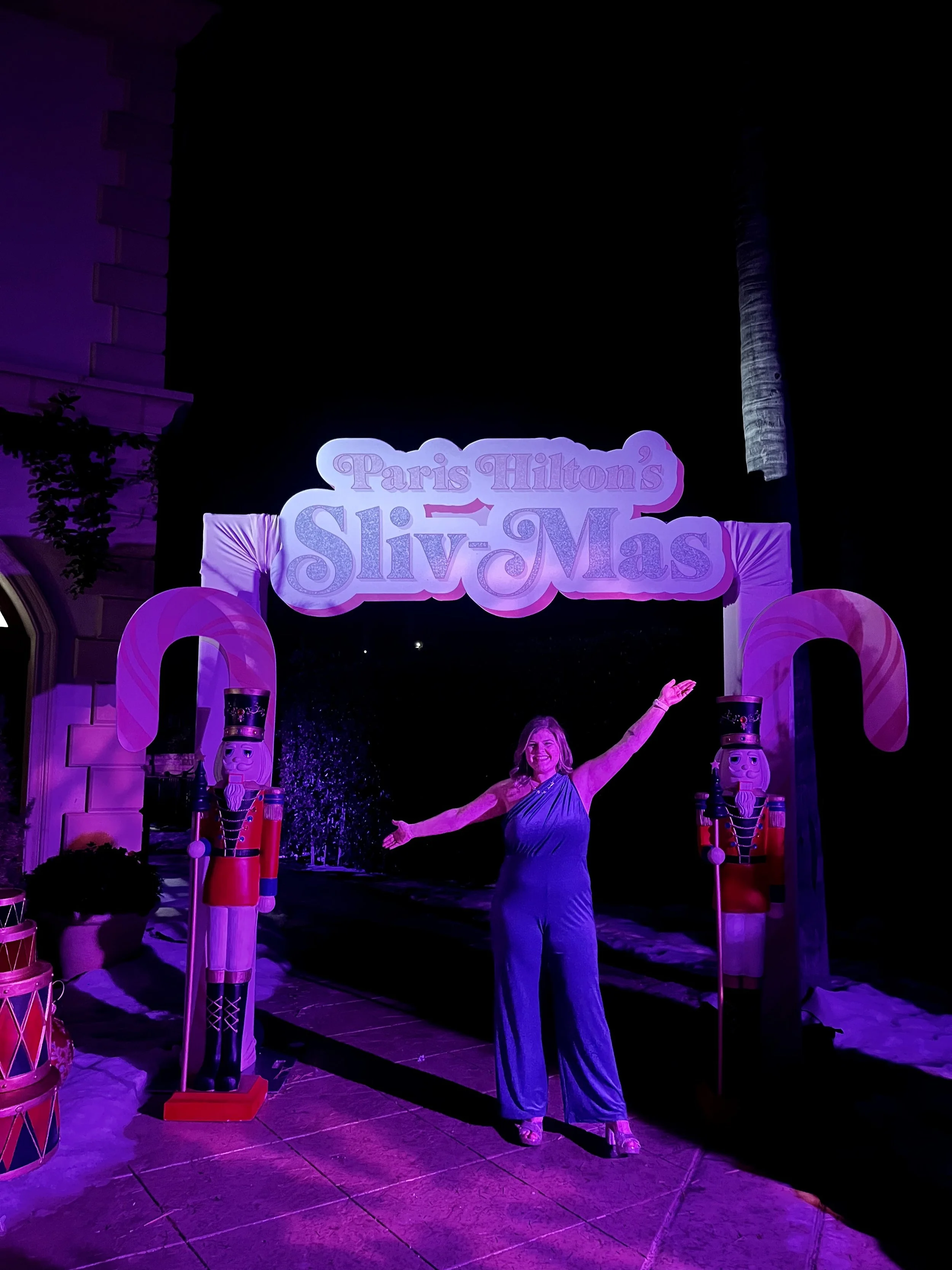 A woman in a blue jumpsuit posing with arms outstretched in front of a sign that reads 'Paris Hilton's Sliv Mas' at night, with purple lighting and decorative nutcracker figures nearby.