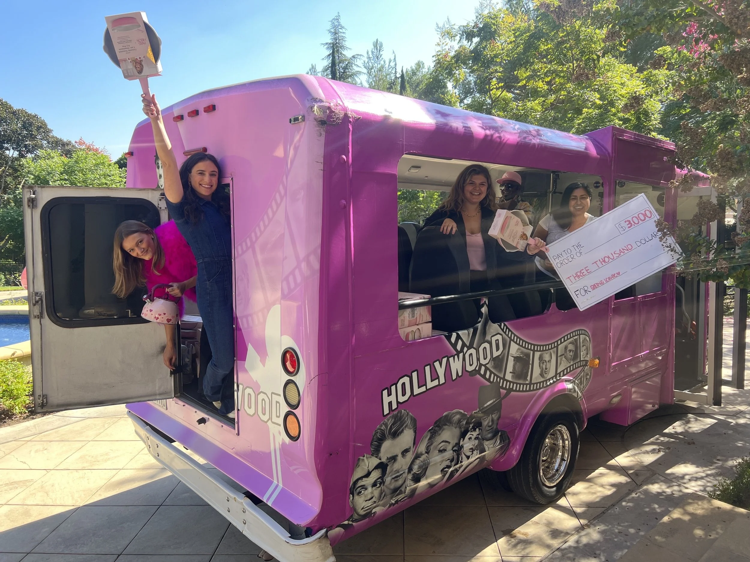 A pink ice cream truck decorated with Hollywood-themed images, including classic movie stars, parked outdoors with trees in the background. Four women and one man are interacting around the truck, with two women holding a large check and ice cream tr
