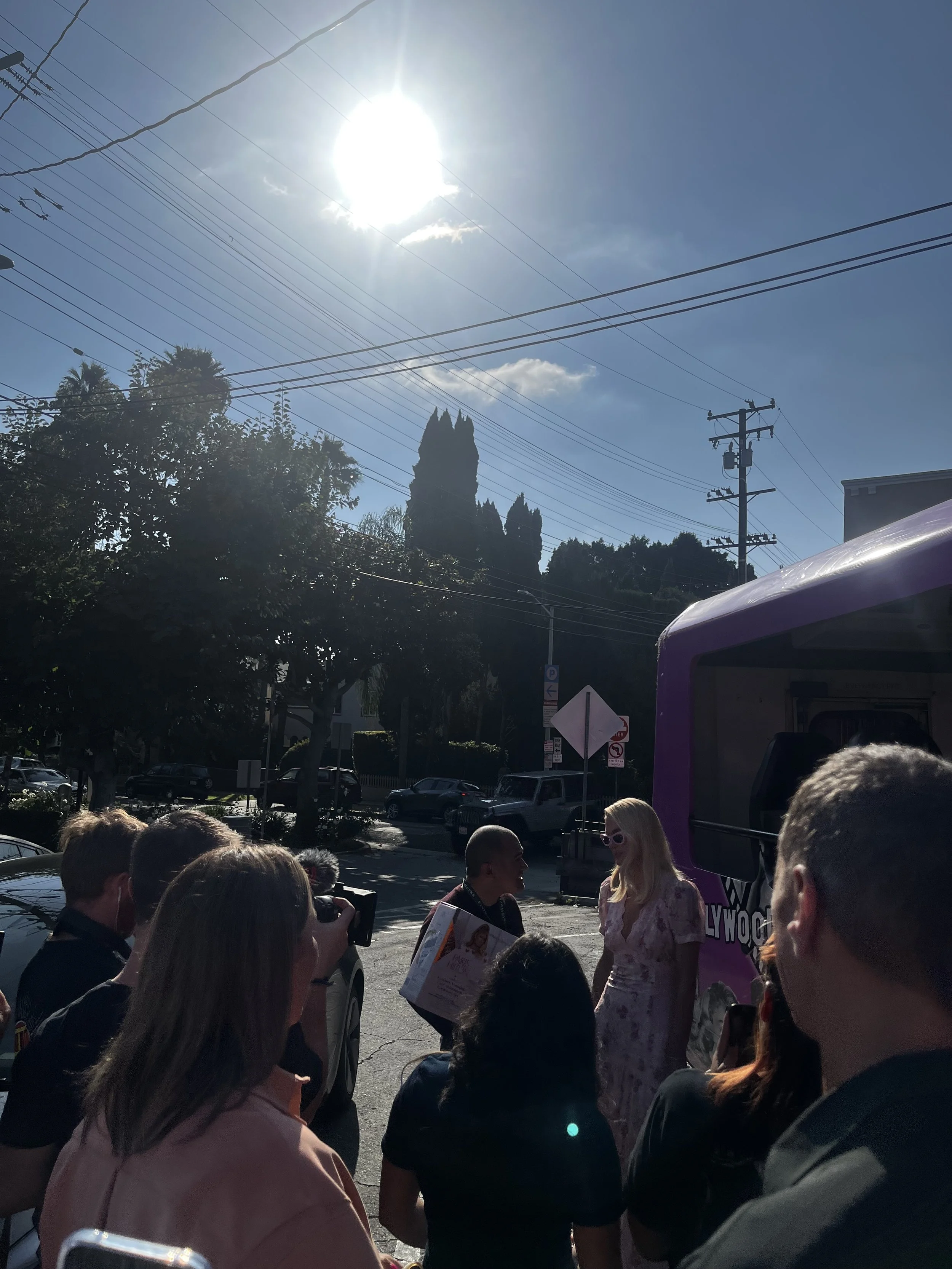 Women and reporters gather outside on sunny day, with a purple vehicle in the background, trees, parked cars, power lines, and a bright sun overhead.