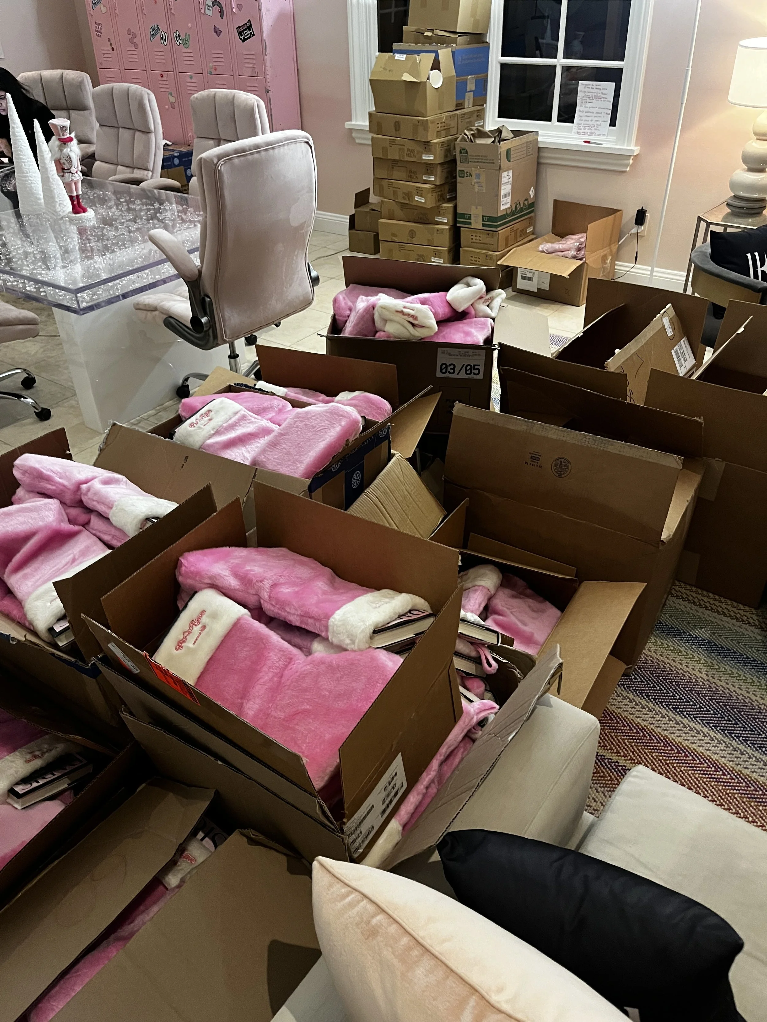 A room with pink lockers, beige chairs around a glass table, and various boxes with pink plush socks and blankets, no people visible.