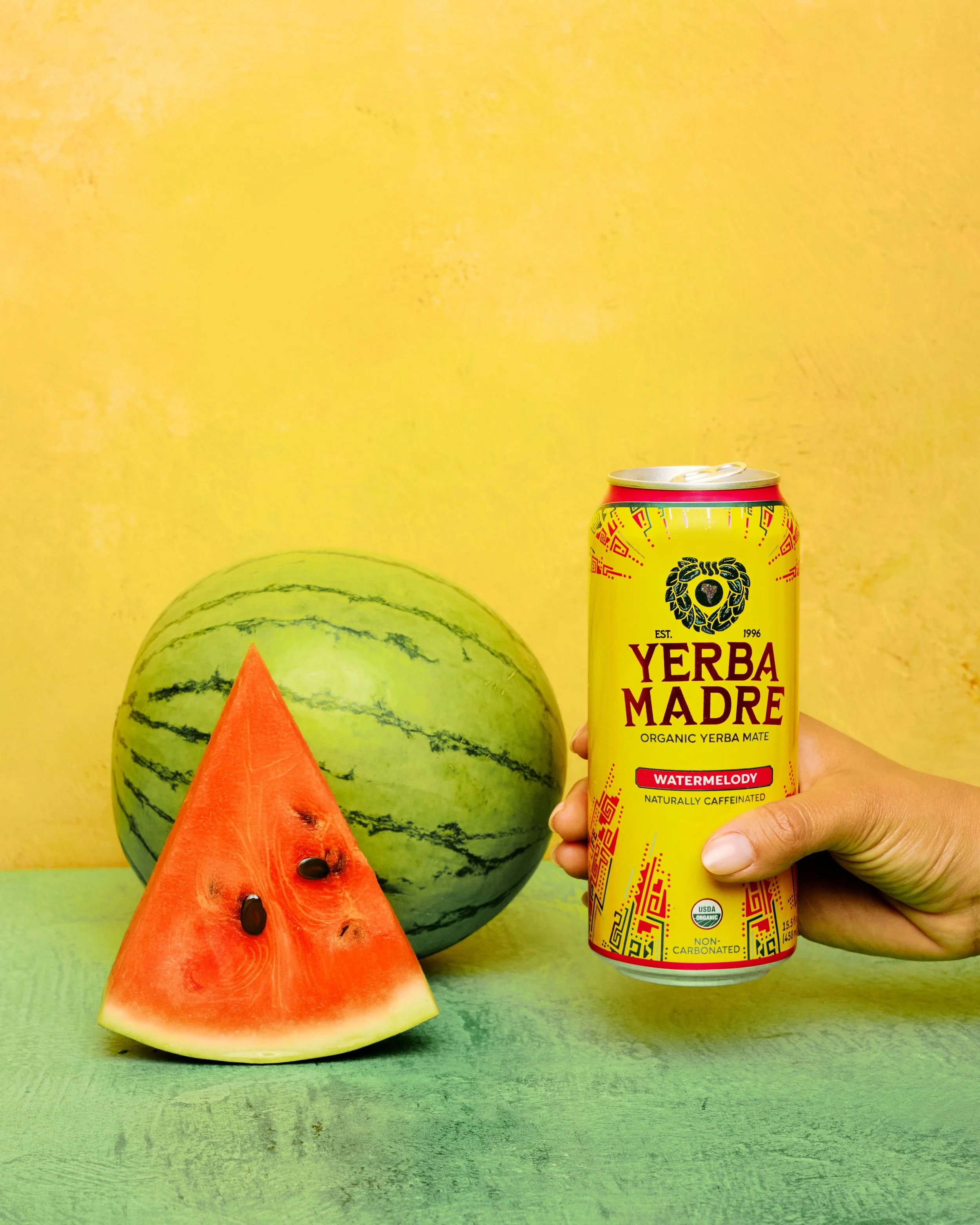 Slice of watermelon with black seeds on green surface, whole watermelon in background, hand holding can of Yerba Madre Watermelon flavored Yerba Mate beverage against yellow wall.