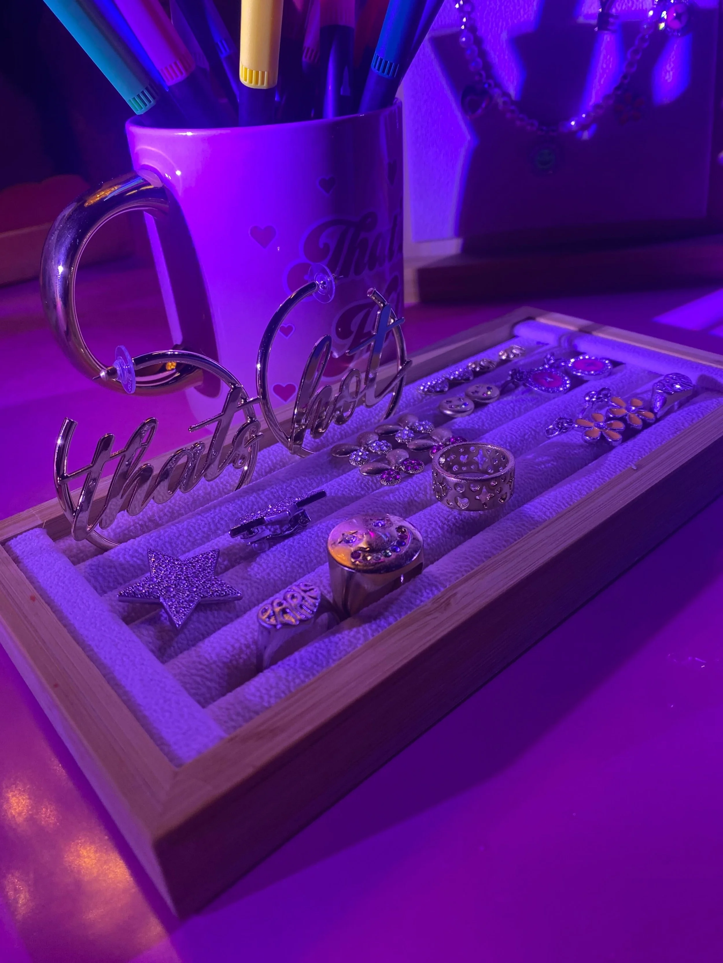 A purple mug with hearts and the words 'That Girl' holds colorful pens. A jewelry box with rings and decorative pins is in front of the mug, with a purple and pink mood lighting.