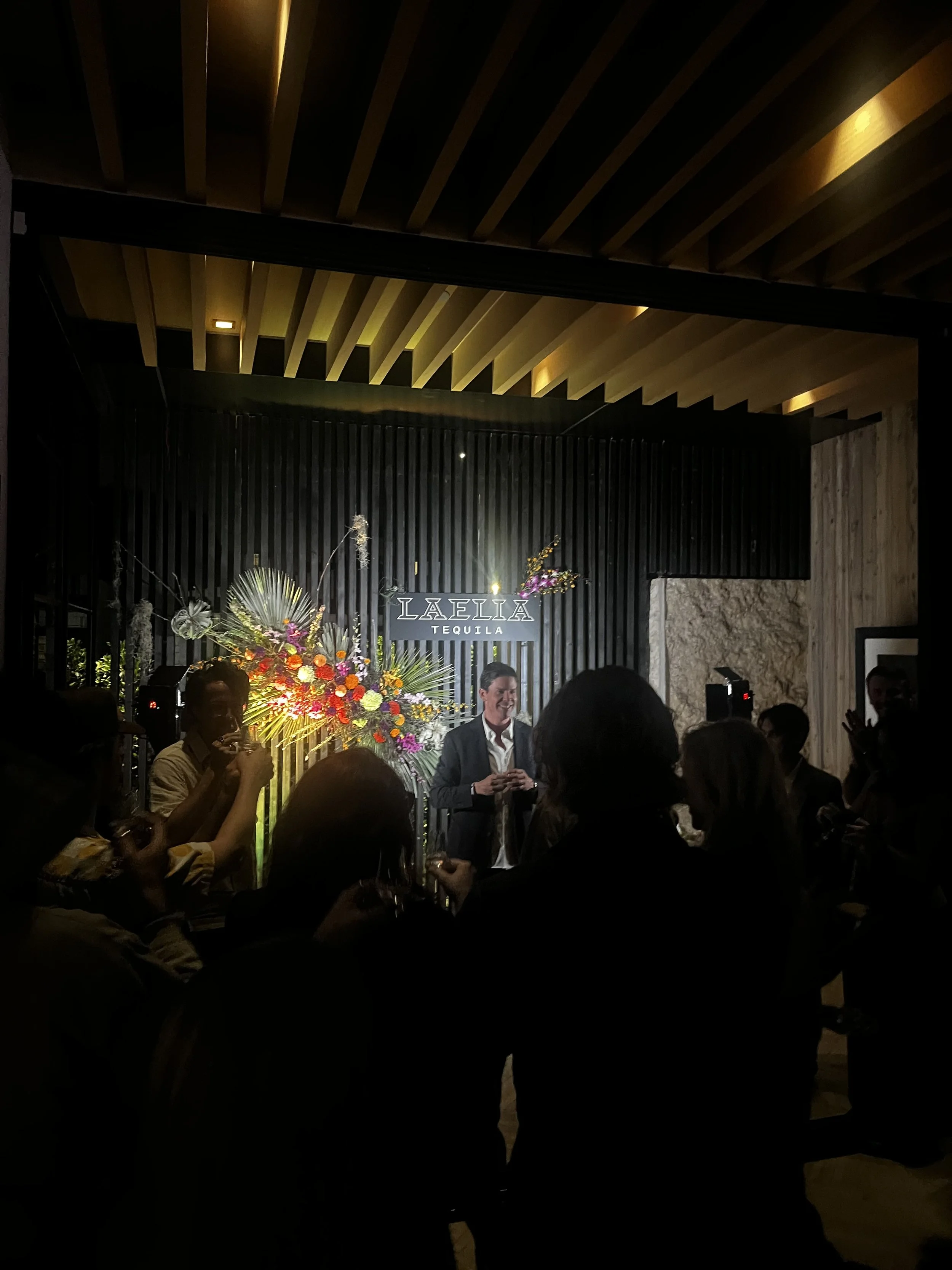 A man in a suit giving a speech at an event with a black backdrop and floral arrangements, with people gathered around, in a modern venue with wooden ceiling beams.