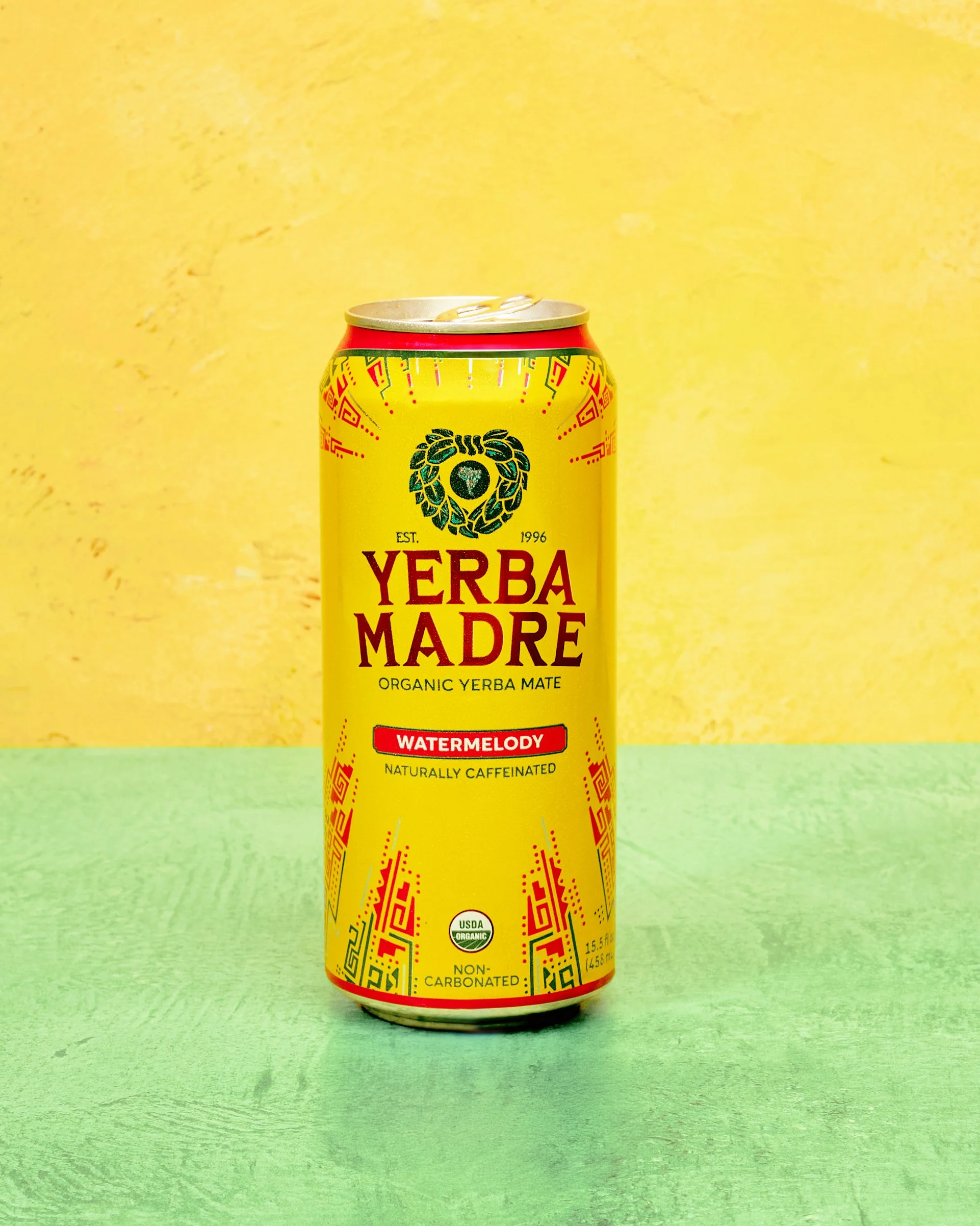 Yellow can of Yerba Madre Organic Yerba Mate with watermelon flavor, placed on a green surface with a yellow background.