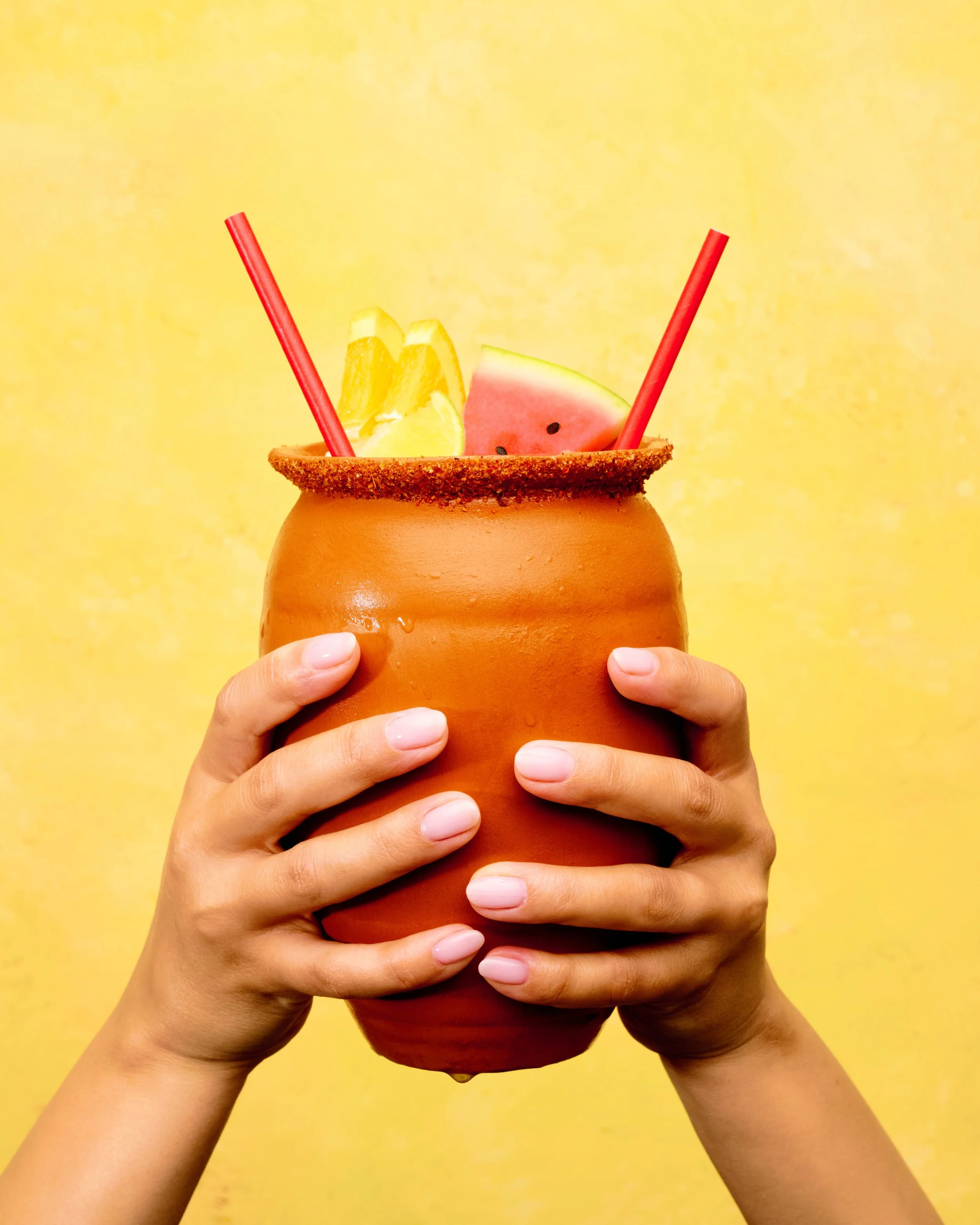 Hands holding a terracotta mug filled with lemon, watermelon, and two red straws against a yellow background.