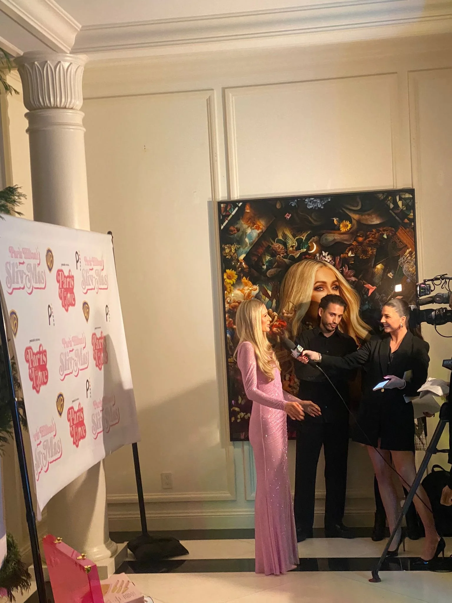 A woman in a long pink dress being interviewed by the media at an event with promotional posters and artwork in the background.