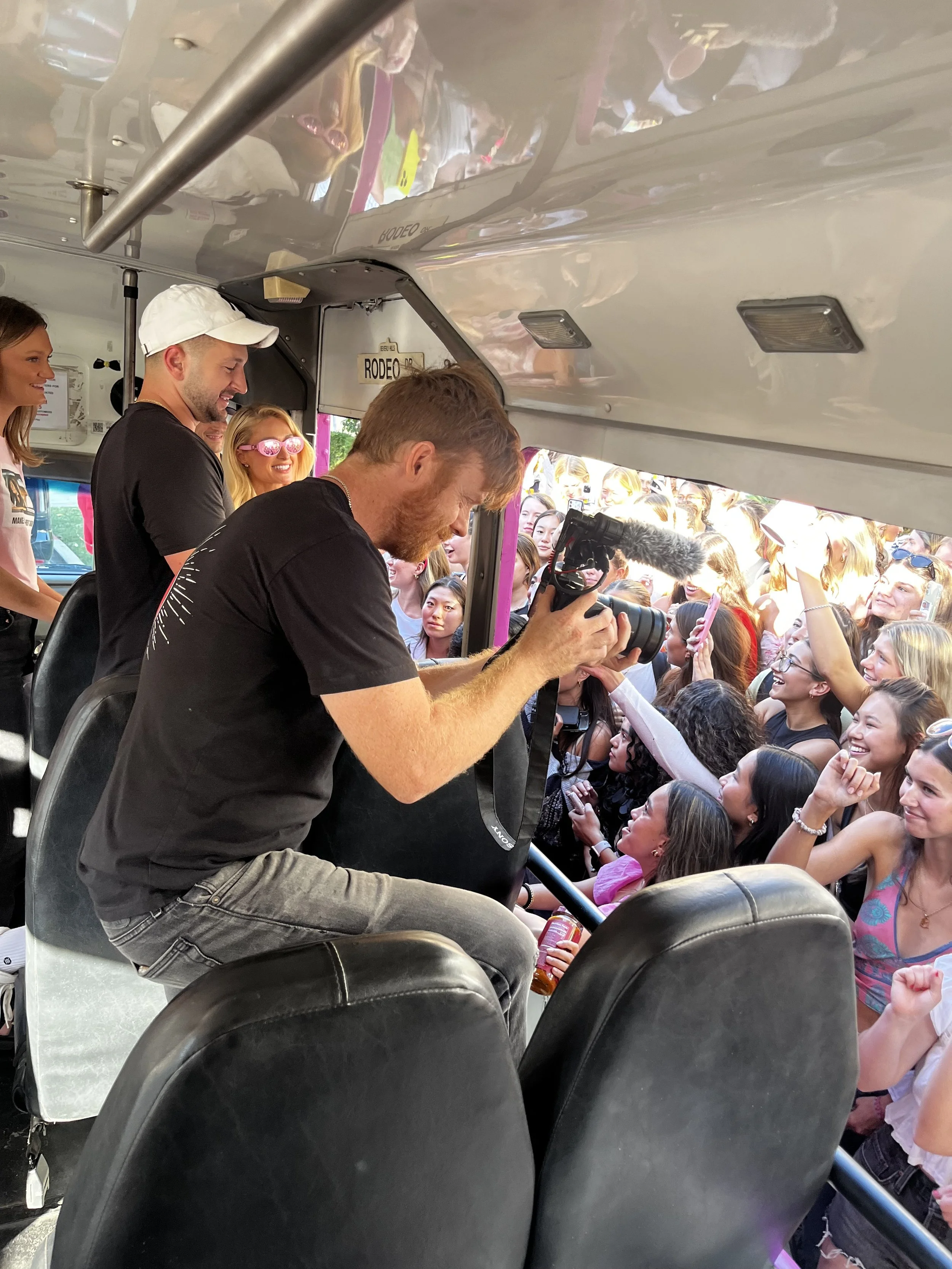 Man taking a photo with a camera inside a bus filled with excited crowd of mostly young women outside the window.