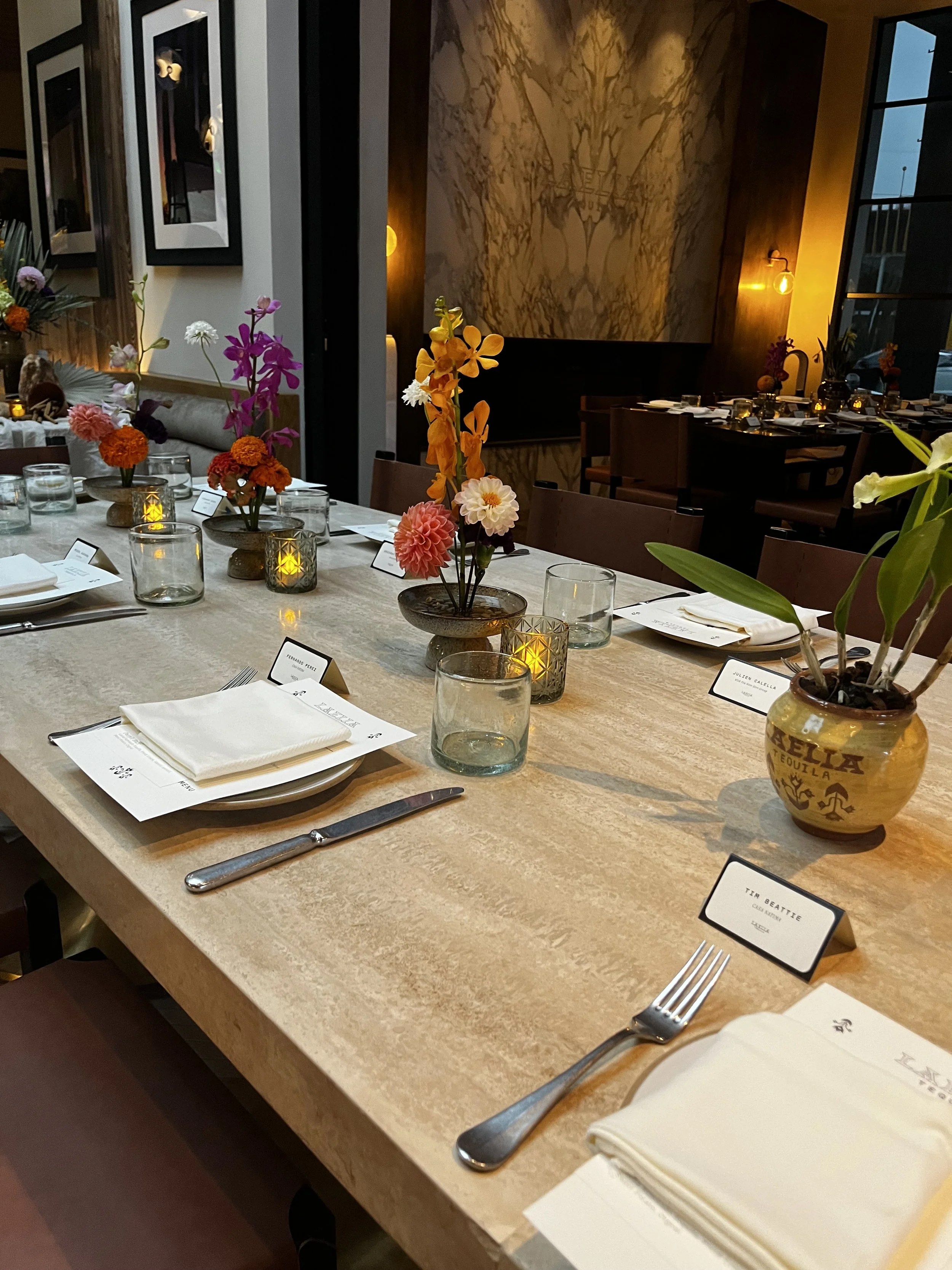 A restaurant table decorated with colorful floral centerpieces, candles, plates, napkins, and silverware. Name cards are placed at each seat, and there is a warm, dimly lit ambiance with large windows.