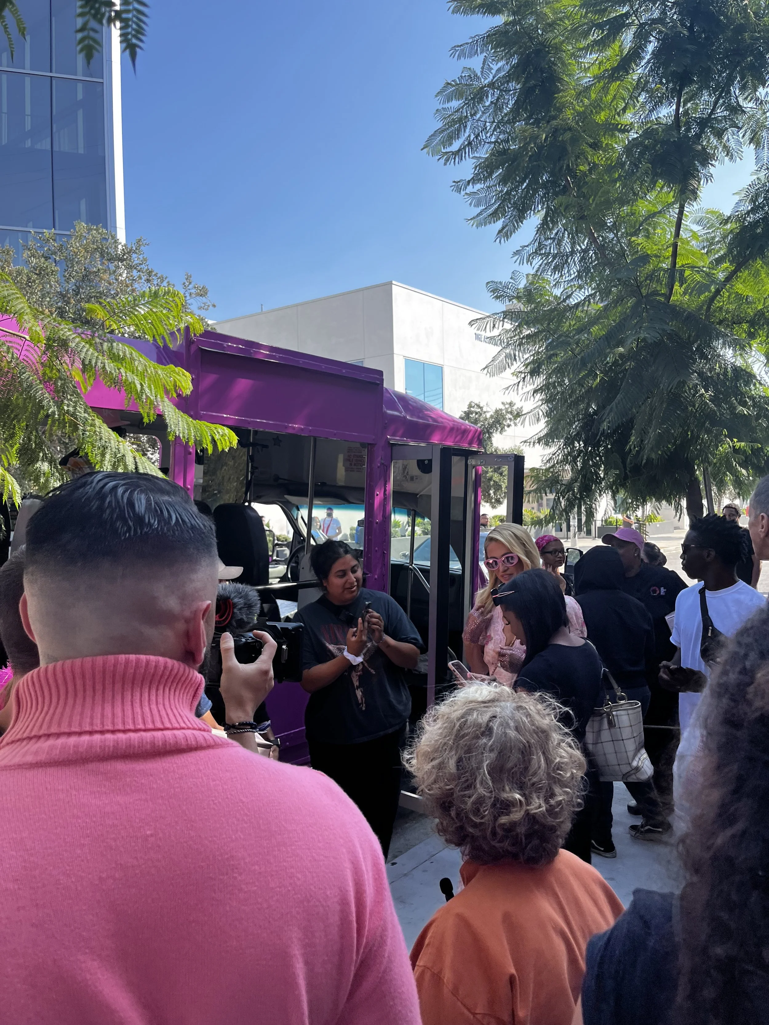 People gathered outside around a purple food truck, some taking photos and talking, with trees and a modern building in the background on a sunny day.