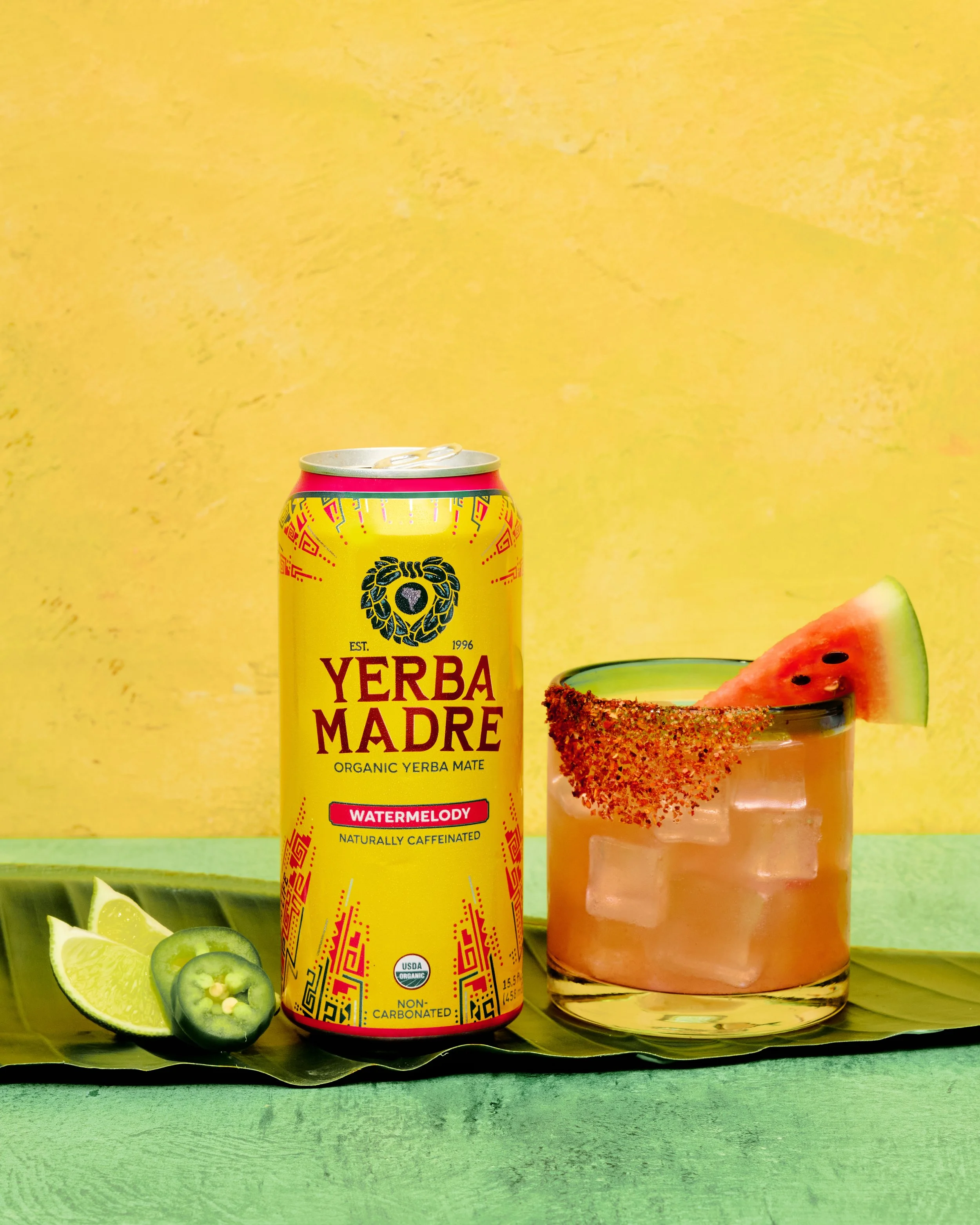 Yellow can of Yerba Madre organíc yerba mate with watermelon flavor, a glass of iced watermelon-flavored drink garnished with a watermelon slice and chili powder rim, lime and jalapeño slices beside the can, on a green surface with a yellow backgroun