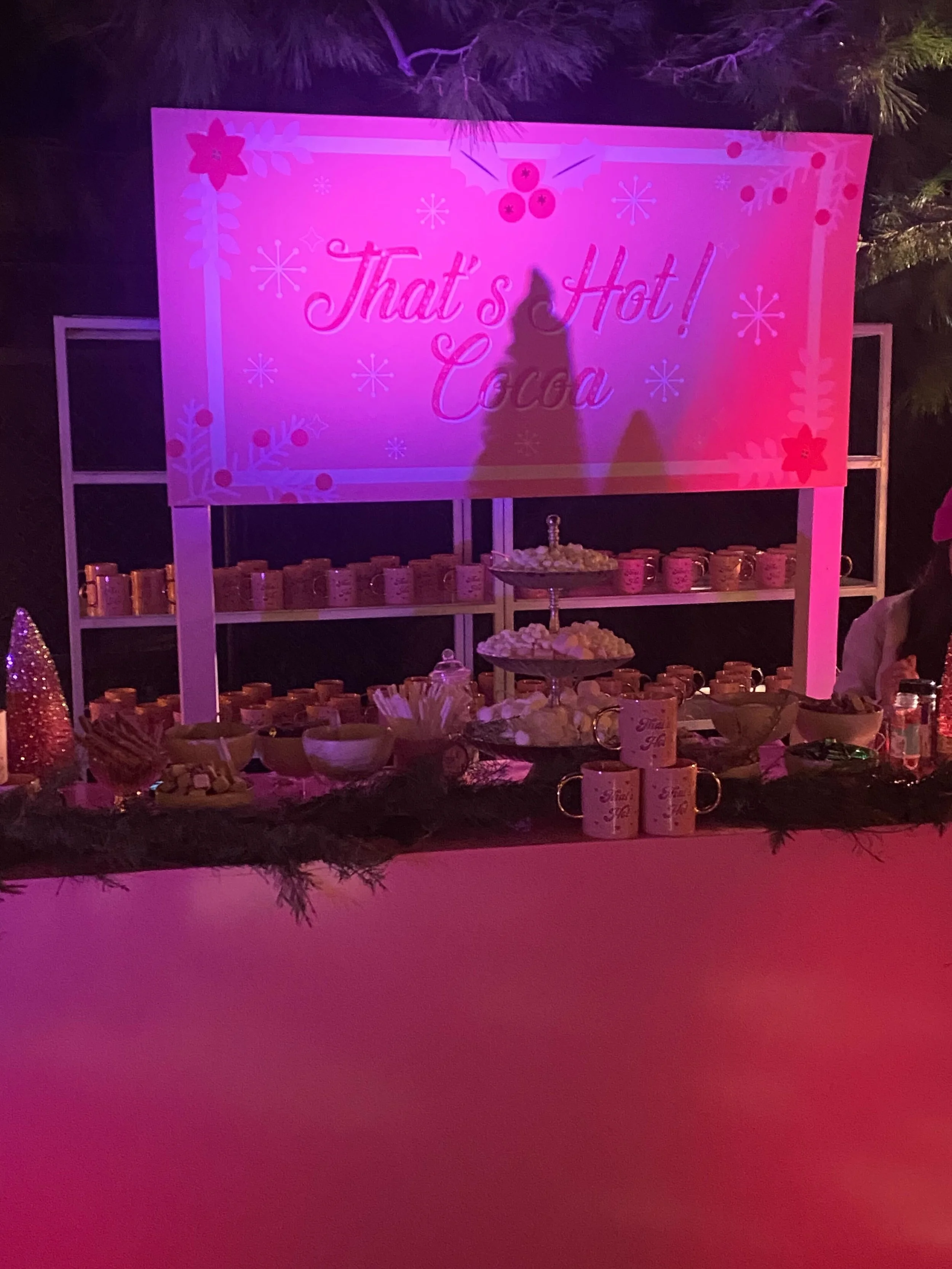 Decorative table with snacks and candies in front of a pink sign that reads 'That's Hot! Coca' at night, decorated for a festive event.