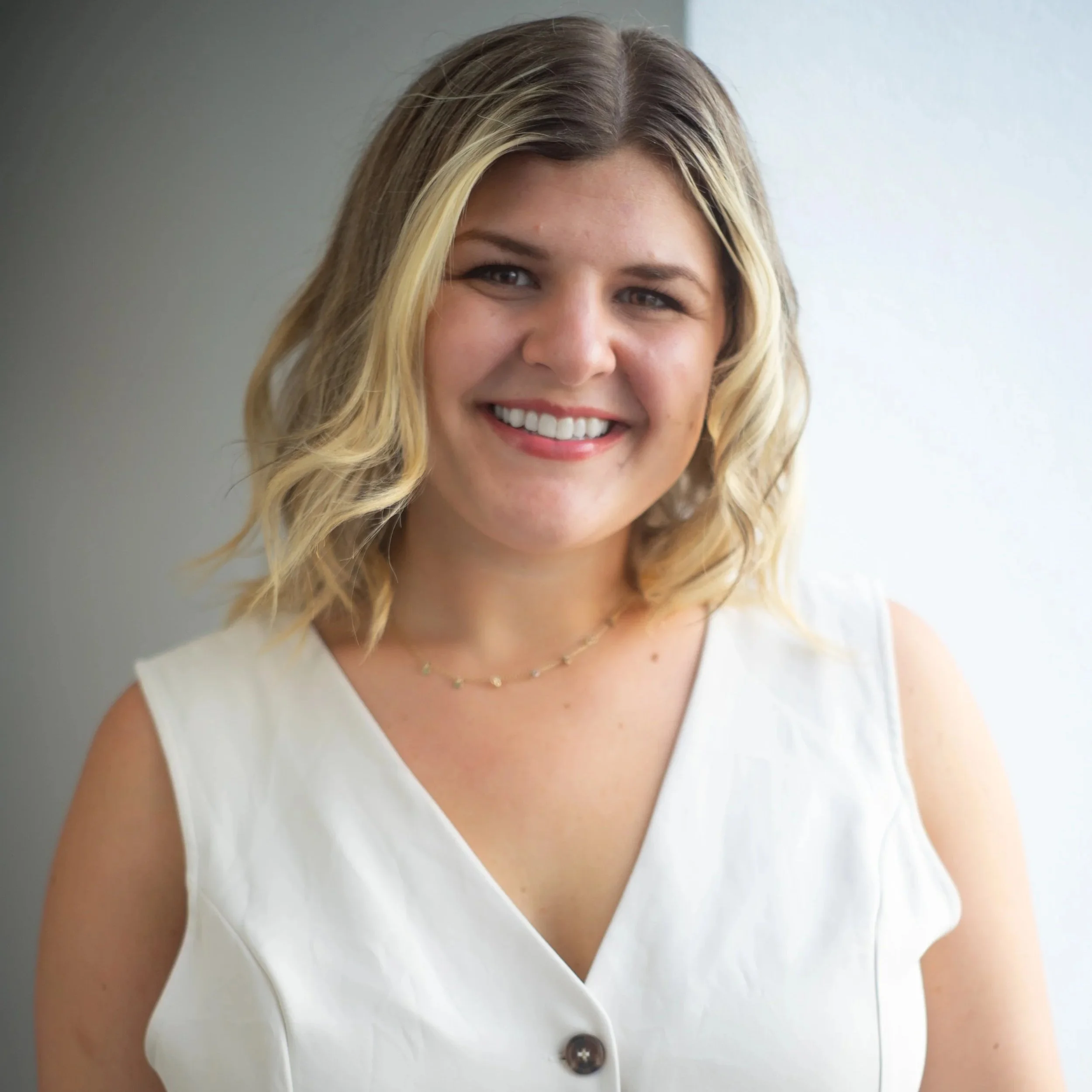 A woman with shoulder-length wavy blonde hair, wearing a sleeveless white top and a delicate necklace, smiling in front of a plain background.
