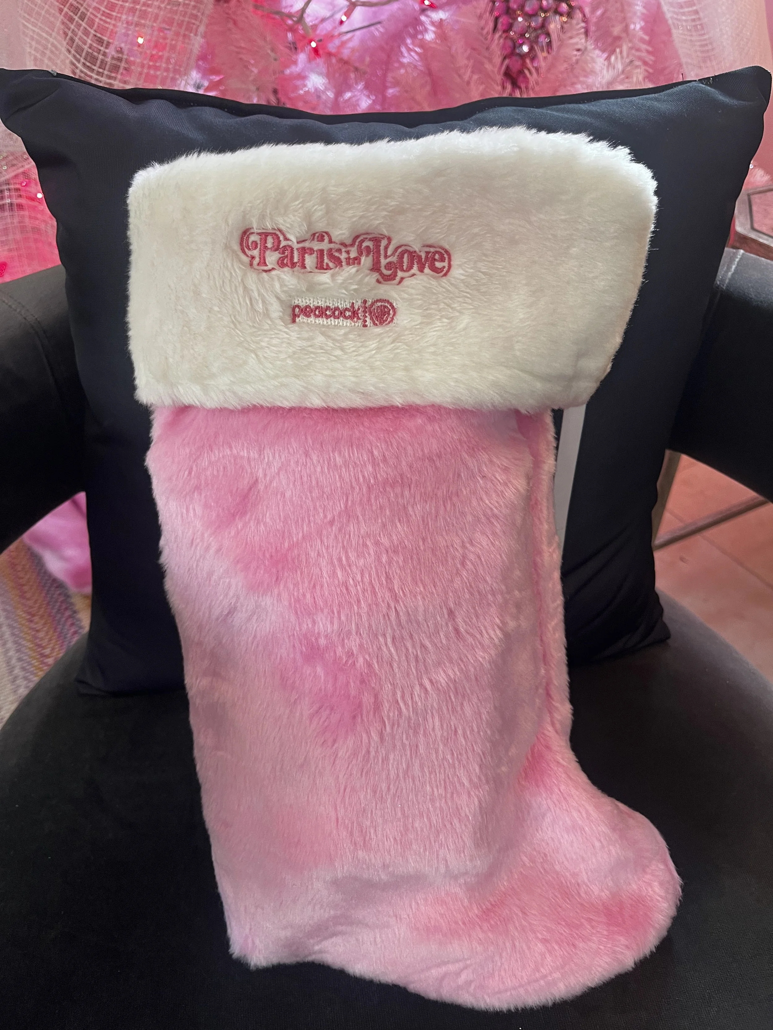 Pink and white Christmas stocking with plush fuzzy texture, hanging on a black cushion. The stocking has embroidery that reads 'Part of Love' and 'peacock'.