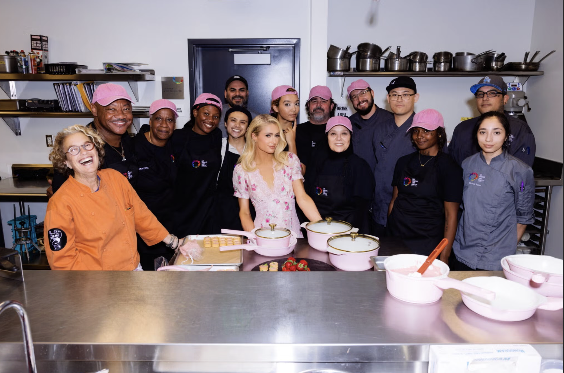 Group of people in a commercial kitchen, wearing chef uniforms and pink hats, gathered around a woman in a floral dress. They are smiling, and the kitchen has stainless steel counters, pink mixing bowls, and cooking utensils on shelves.
